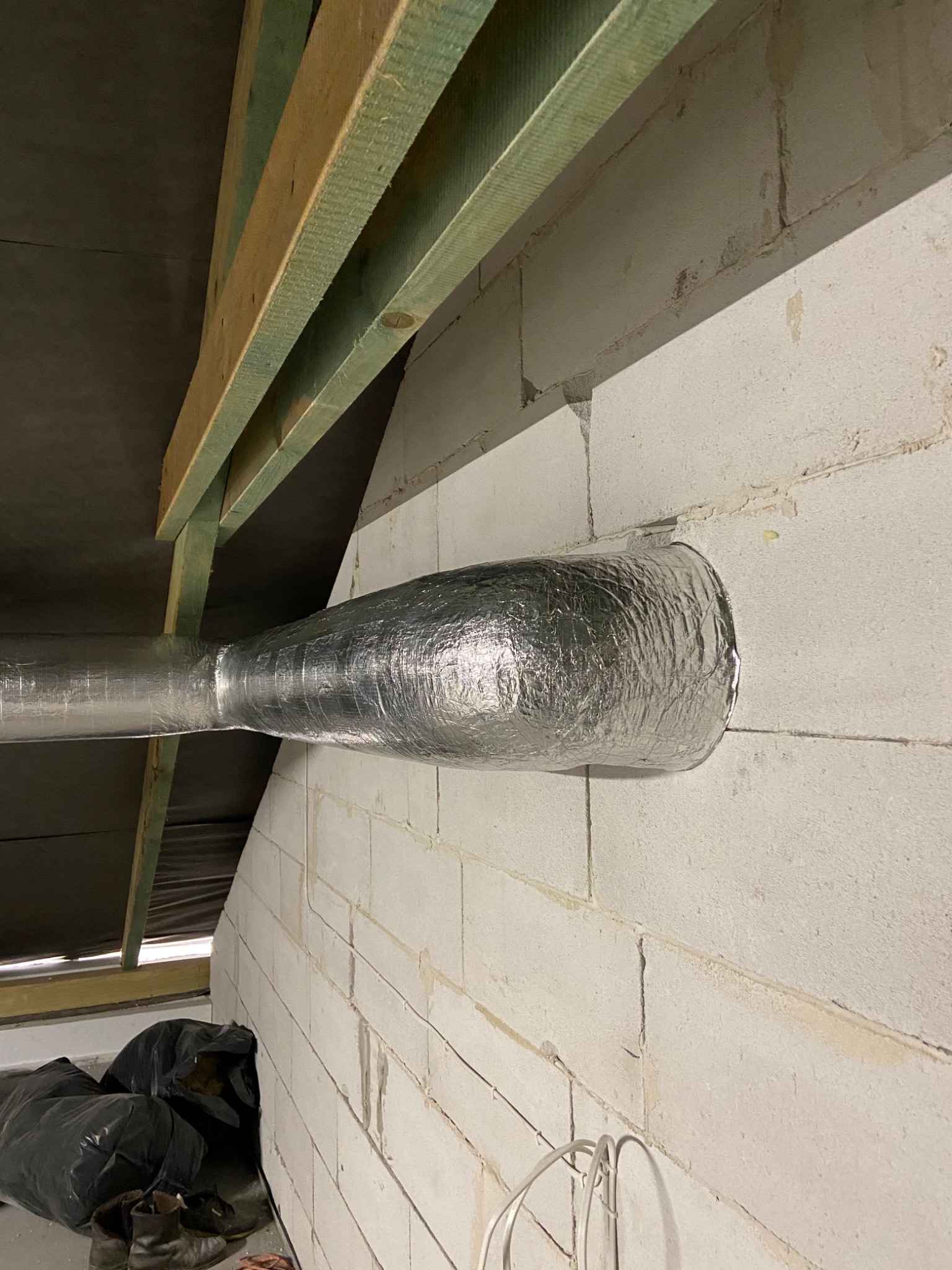 Izolowany przewód wentylacyjny przechodzący przez ścianę z betonu komórkowego, widoczne drewniane elementy konstrukcji dachu oraz czarne worki w tle.