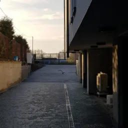 Ułożona, ciemnoszara kostka brukowa na podjeździe, z widocznym odpływem liniowym po prawej stronie, prowadząca w kierunku bramy wjazdowej. Budynek po prawej stronie w cieniu, z widocznymi...