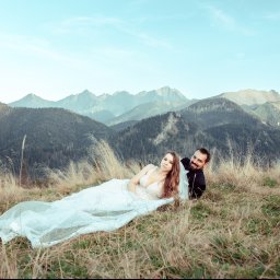 BigStories - Para młoda leżąca na trawie w górach, ona w sukni ślubnej z koronkowymi zdobieniami, on w czarnym garniturze, w tle malowniczy krajobraz górski pod błękitnym niebem.