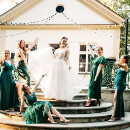 BigStories - Panna młoda w koronkowej sukni i druhny w szmaragdowych sukniach pozują na schodach przed budynkiem z girlandami świetlnymi.