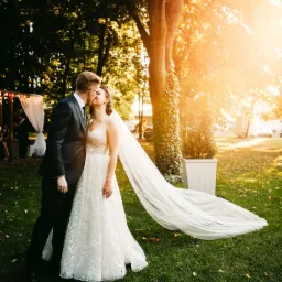 Pocałunek nowożeńców w plenerze, ona w koronkowej sukni z długim welonem, on w ciemnym garniturze, złote światło słoneczne przebija przez drzewa.