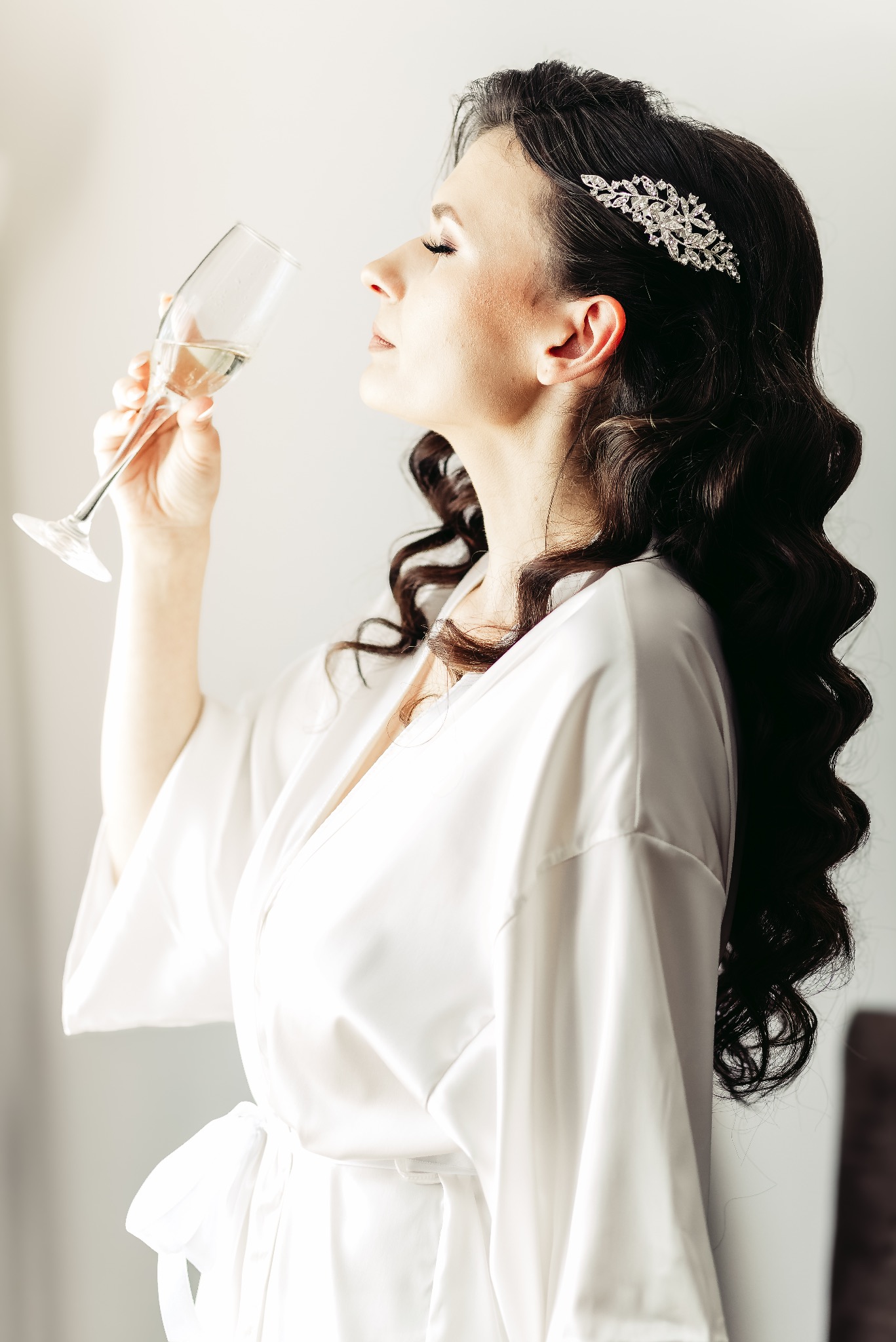 Panna młoda w białym szlafroku, z upiętymi ciemnymi włosami ozdobionymi błyszczącą spinką, pije szampana przed ślubem.