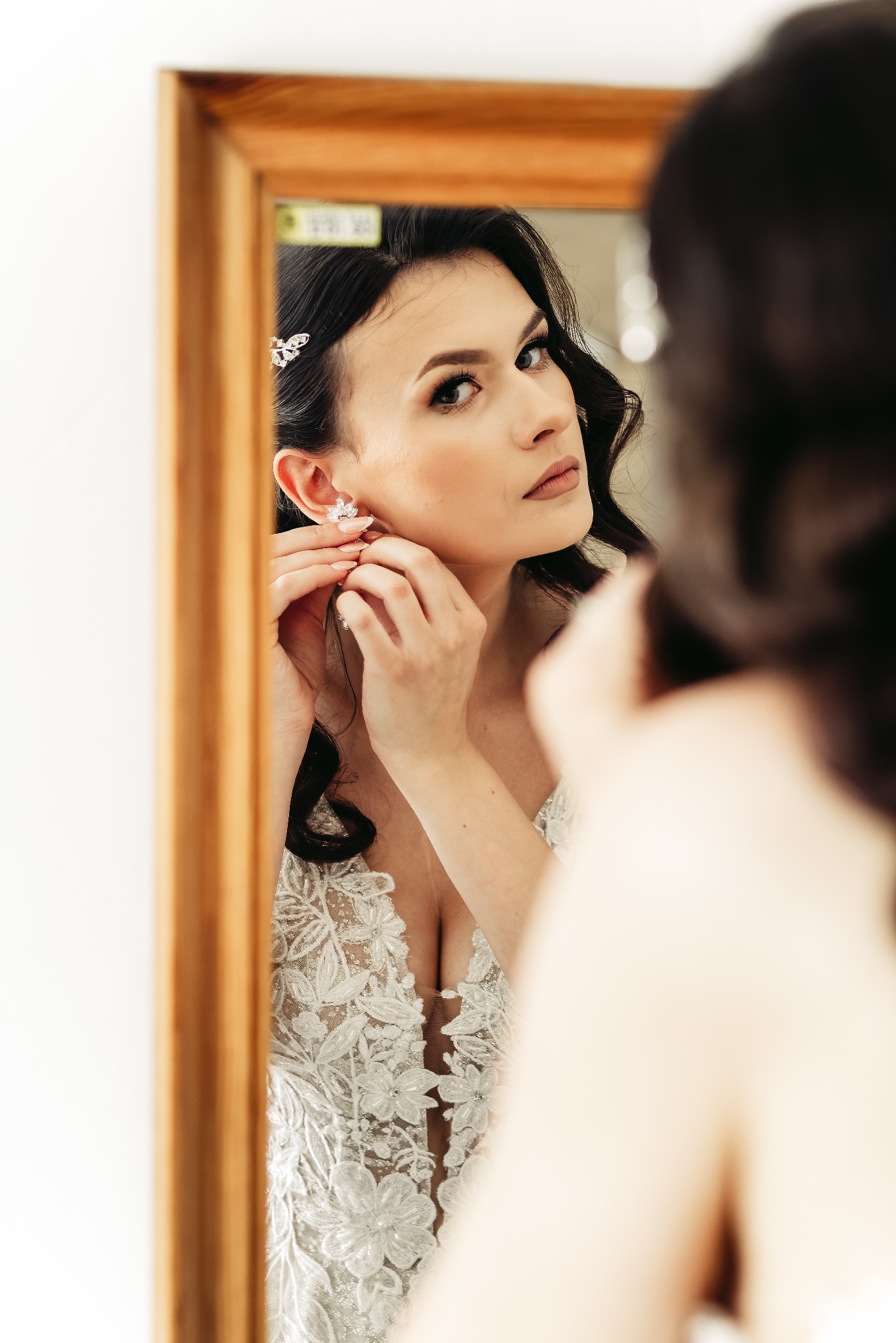 Panna młoda z ciemnymi włosami, w białej sukni z koronkowymi aplikacjami, zakłada kolczyk przed lustrem w drewnianej ramie, przygotowując się do ślubu.