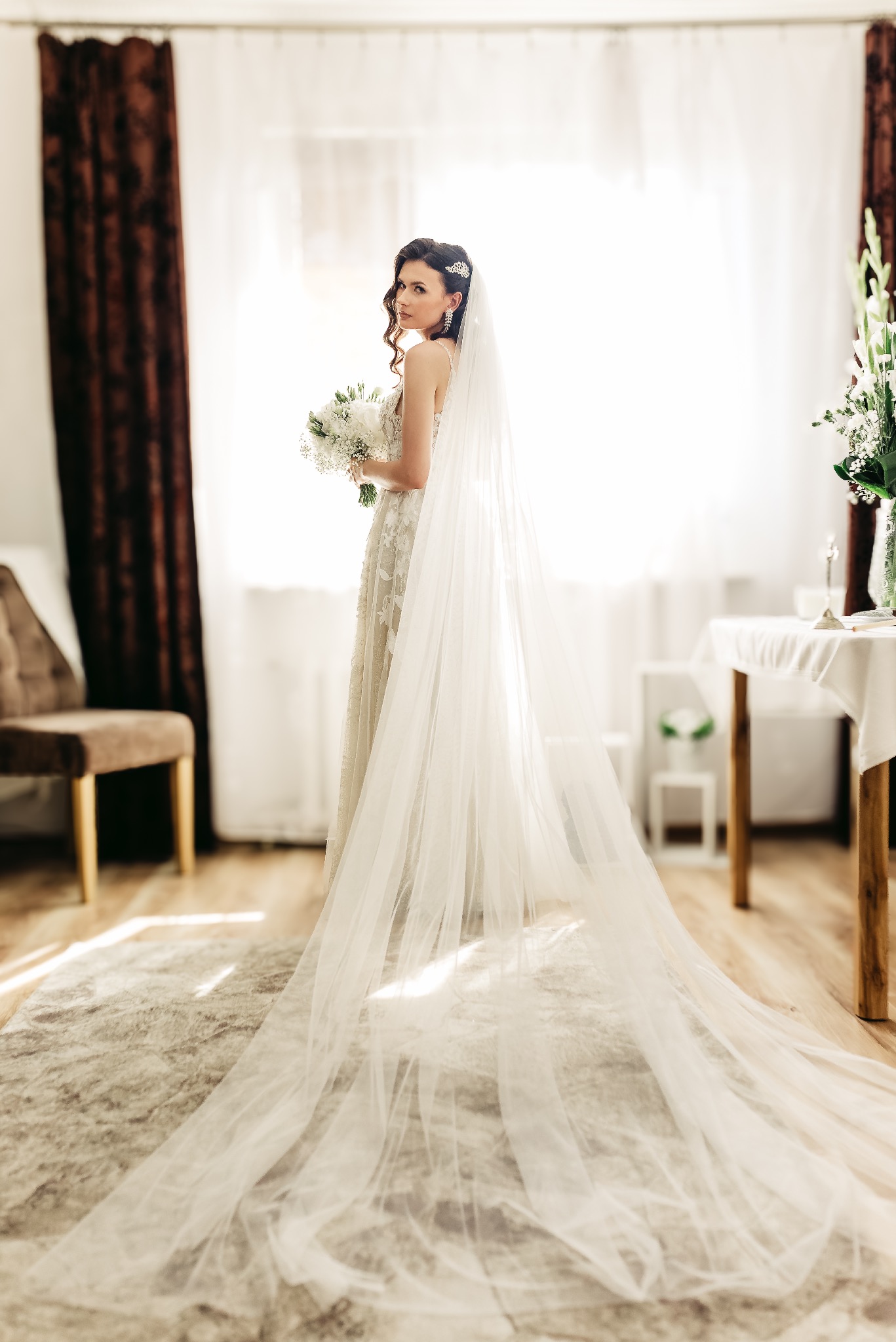 Panna młoda w koronkowej sukni i długim welonie, trzymająca bukiet białych kwiatów, spogląda przez ramię w jasnym wnętrzu z brązowymi zasłonami.