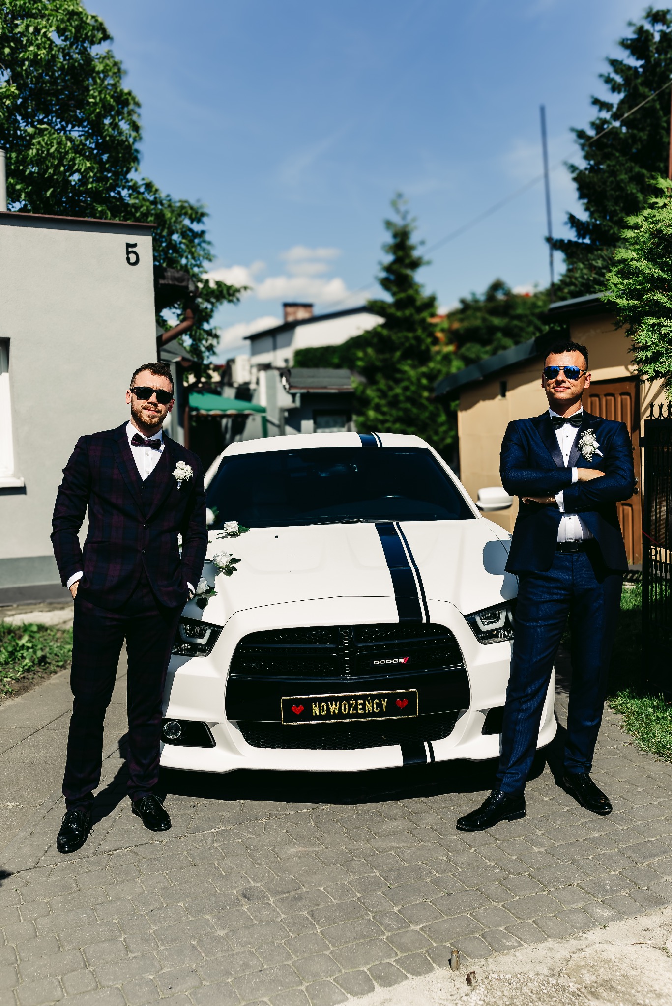 Dwóch elegancko ubranych mężczyzn, jeden w bordowym garniturze w kratę, drugi w granatowym, pozuje obok białego samochodu Dodge z czarnym pasem i tablicą rejestracyjną 'Nowożeńcy', kwiatowe...