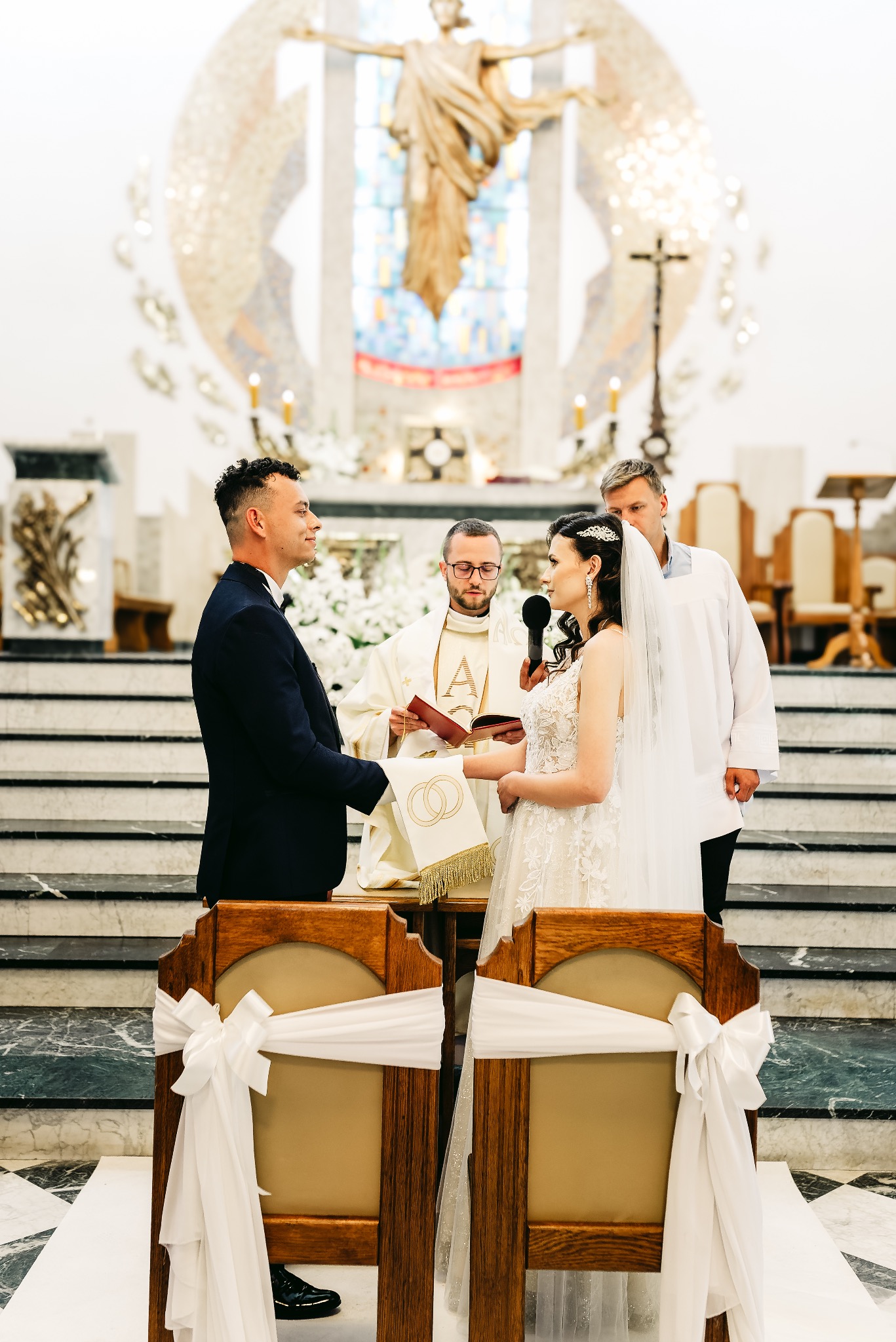 Para młoda podczas ceremonii ślubnej w kościele, z widocznym księdzem, świadkiem i dekoracjami kwiatowymi w tle. W pierwszym planie krzesła z białymi kokardami.