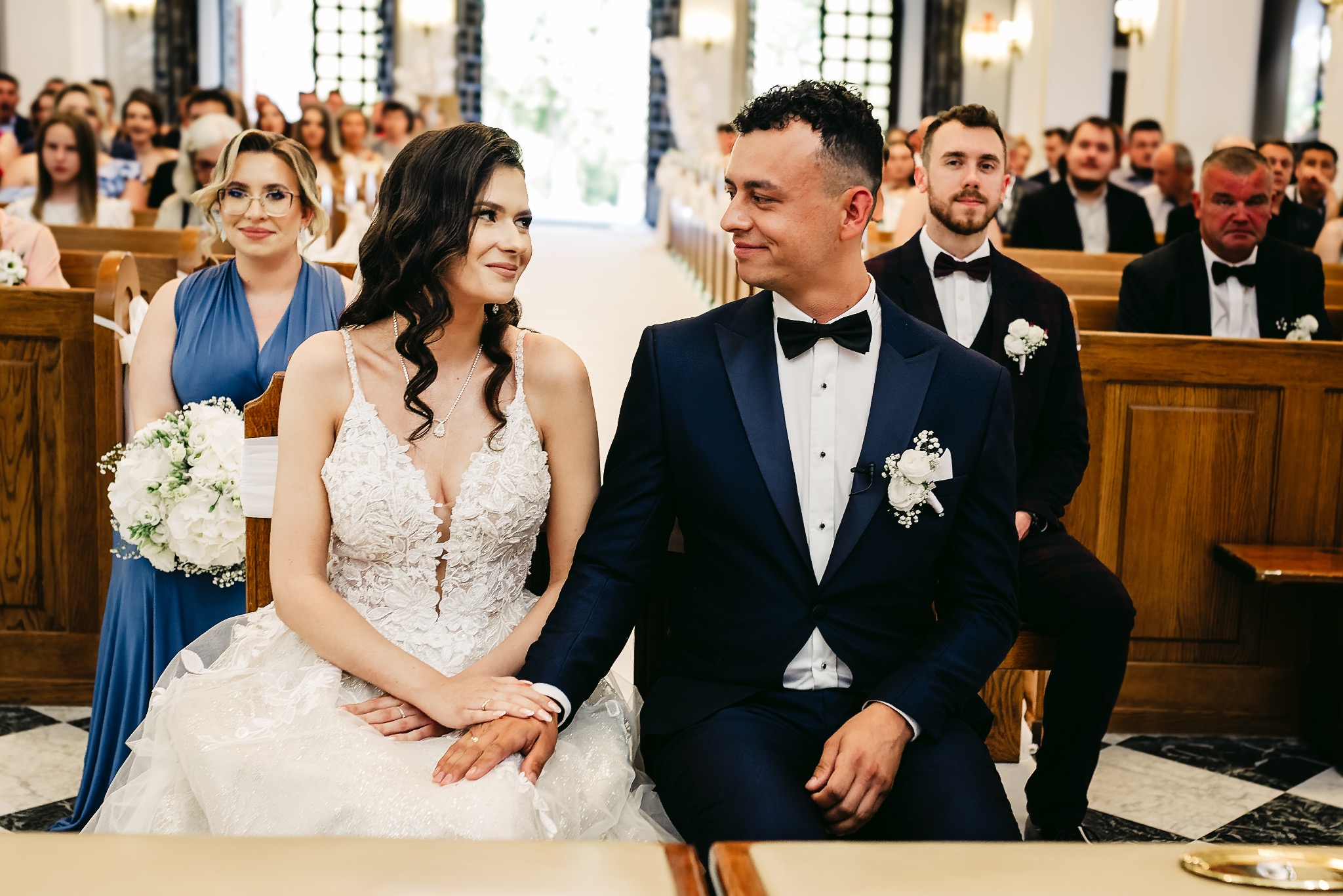 Para młoda siedzi obok siebie w kościele, spoglądając na siebie z uśmiechem, w tle goście weselni.