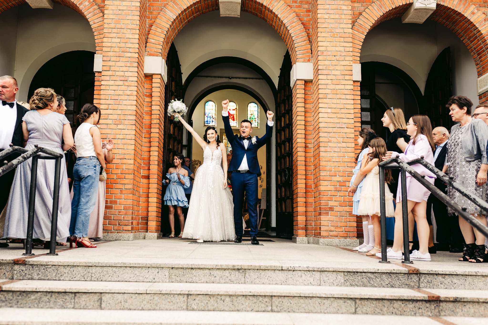 Para nowożeńców w strojach ślubnych wychodzi z kościoła z uniesionymi rękami, otoczona gośćmi weselnymi na tle ceglanej fasady z łukowymi wejściami.