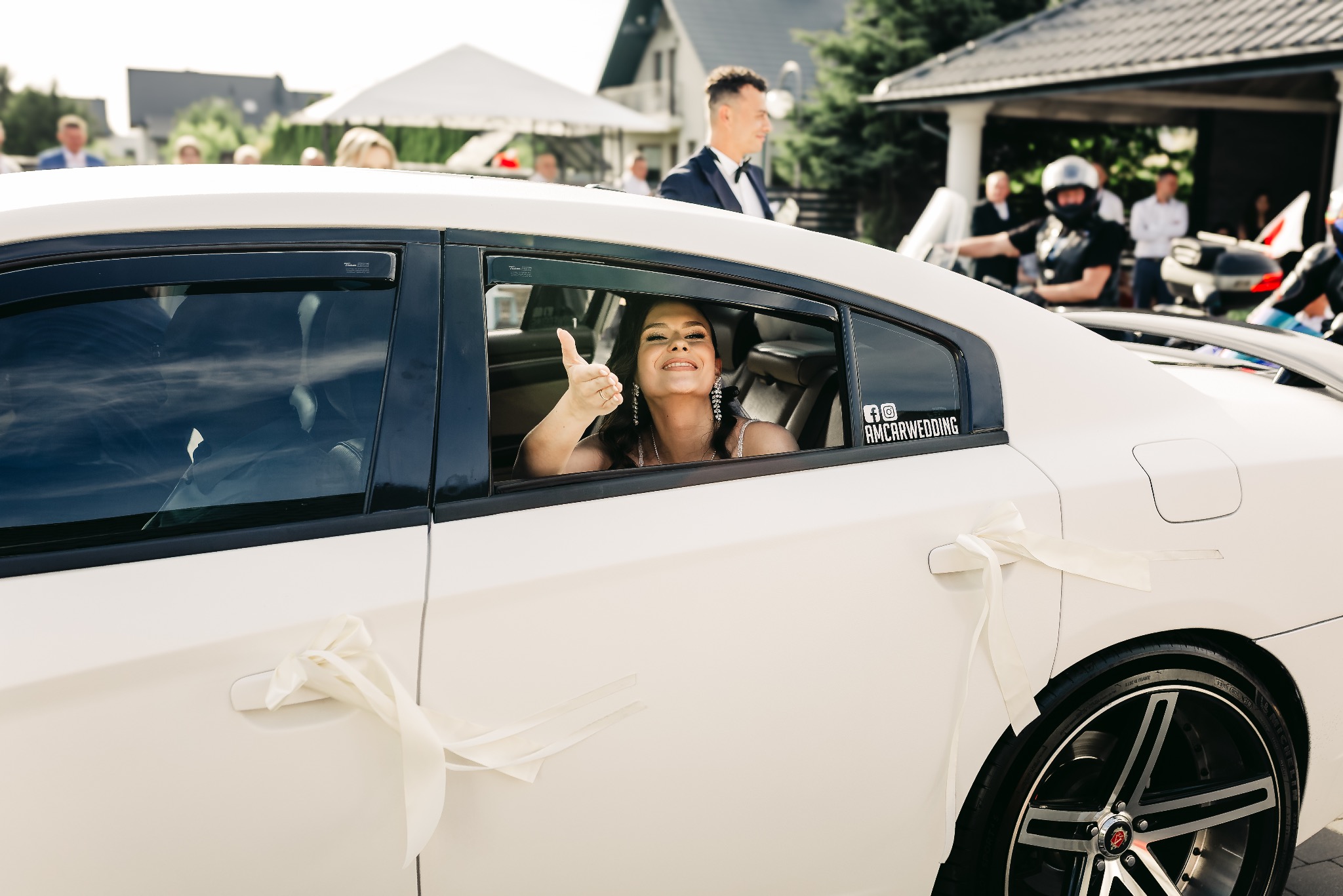 Uśmiechnięta panna młoda w białej sukni, wystawia kciuk w górę z tylnego siedzenia białego samochodu ślubnego ozdobionego wstążkami, w tle goście weselni i budynki.