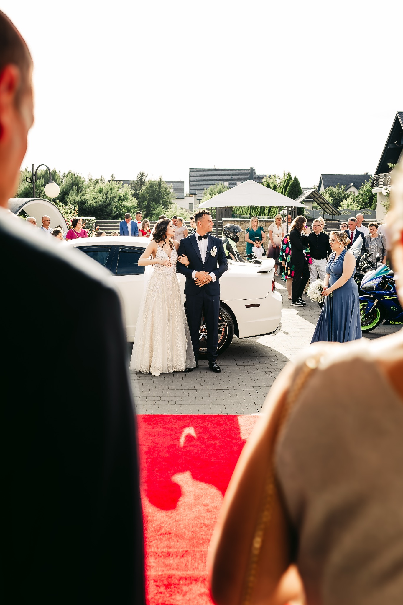 Para młoda wychodzi z białej limuzyny na czerwony dywan, w tle goście weselni i niebieski motocykl.