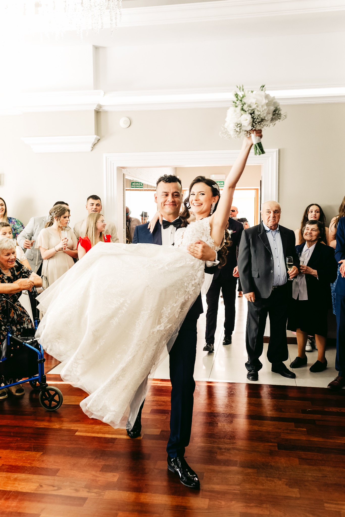 Pan młody trzyma na rękach uśmiechniętą pannę młodą z bukietem, goście weselni w tle, parkiet z ciemnego drewna.