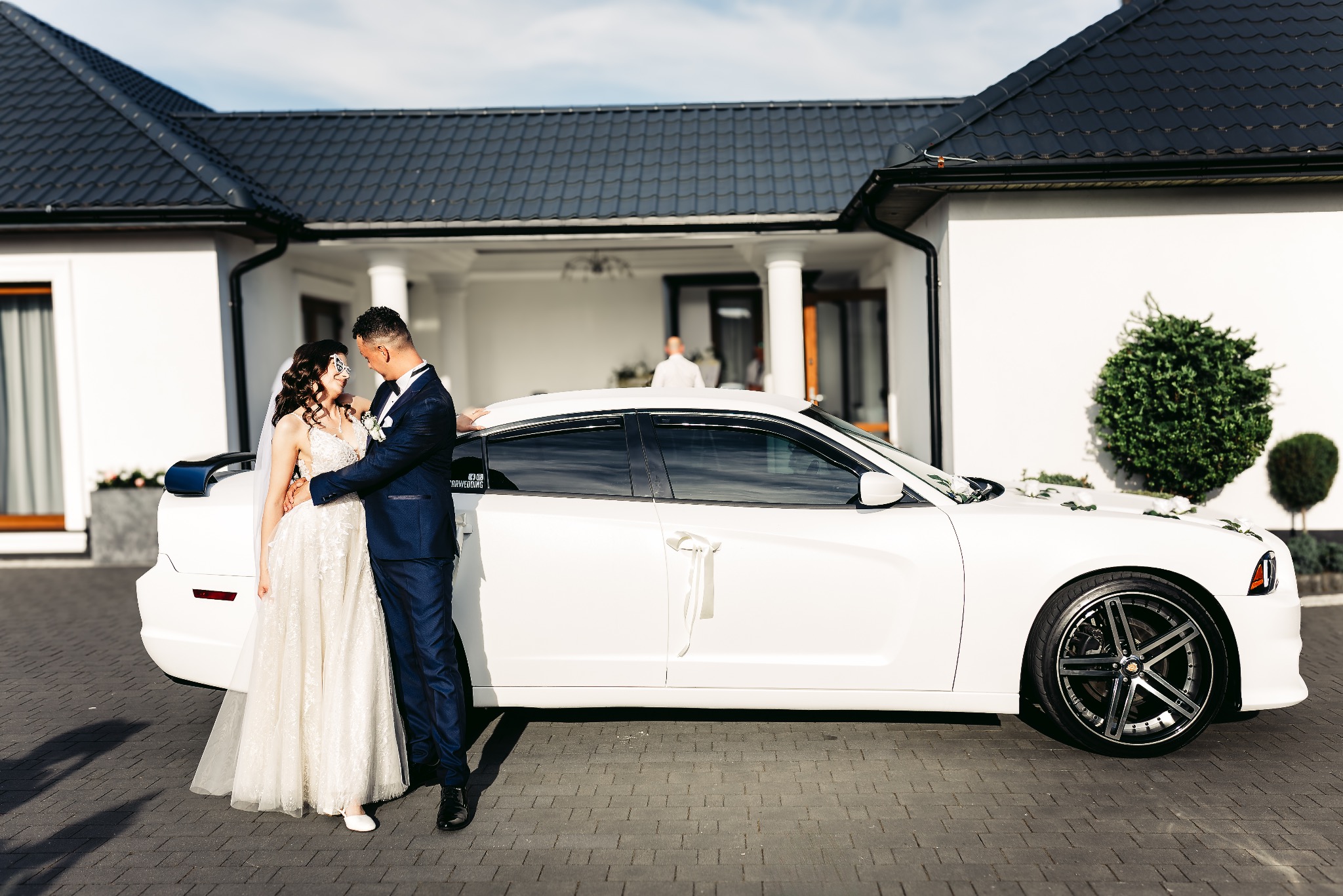 Para młoda w objęciach przy białym, udekorowanym samochodzie na tle nowoczesnego domu z ciemnym dachem.