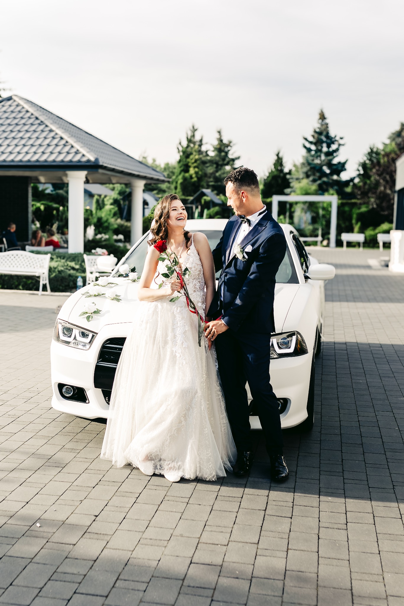 Uśmiechnięta panna młoda w koronkowej sukni trzyma czerwoną różę, opierając się o biały samochód ozdobiony białymi różami, obok pan młody w granatowym garniturze z muchą, na tle budynku z kolumnami...