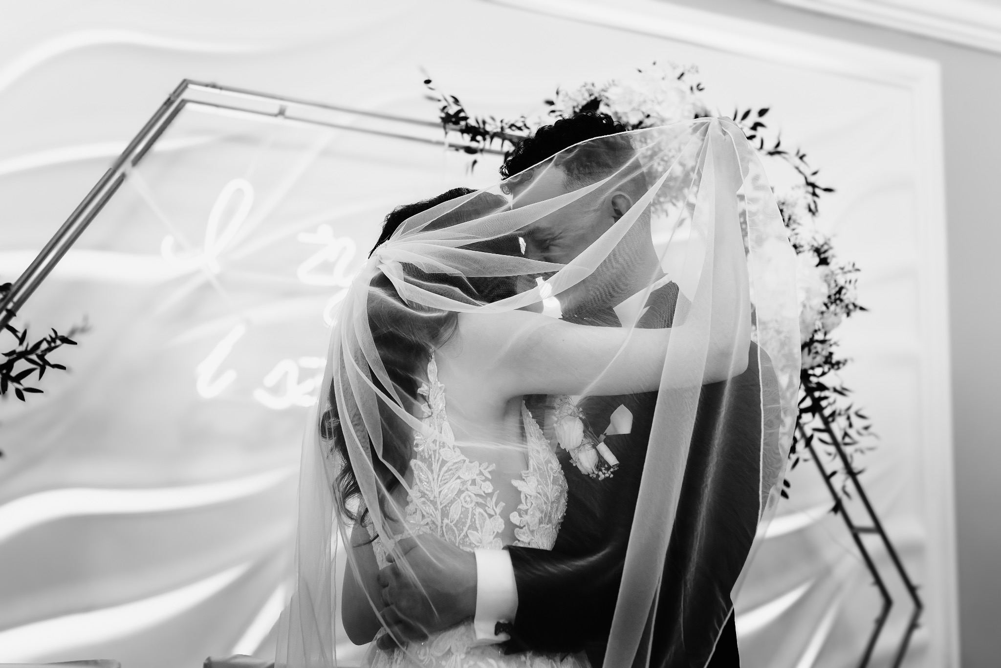 Czarno-białe zdjęcie obejmującej się pary młodej pod welonem, z dekoracją kwiatową i neonowym napisem w tle.