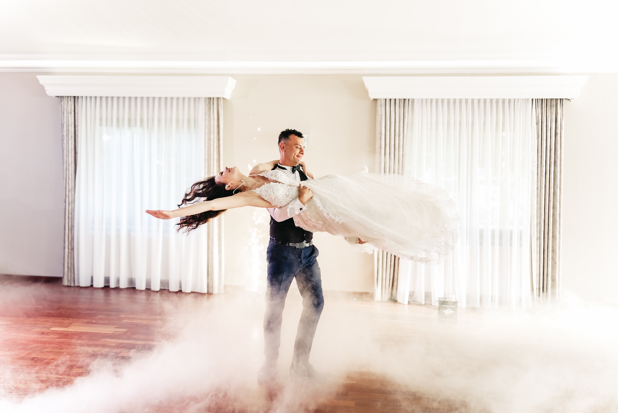 Pan młody trzyma pannę młodą w ramionach podczas pierwszego tańca w otoczeniu sztucznego dymu na parkiecie.