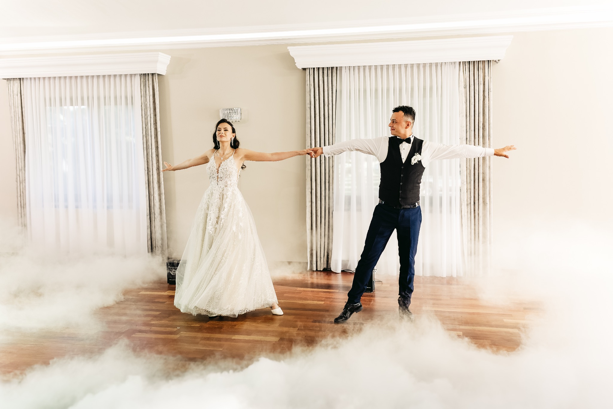 Para młoda, Panna Młoda w koronkowej sukni i Pan Młody w czarnej kamizelce, tańczą w oparach białego dymu na parkiecie w jasnym wnętrzu.