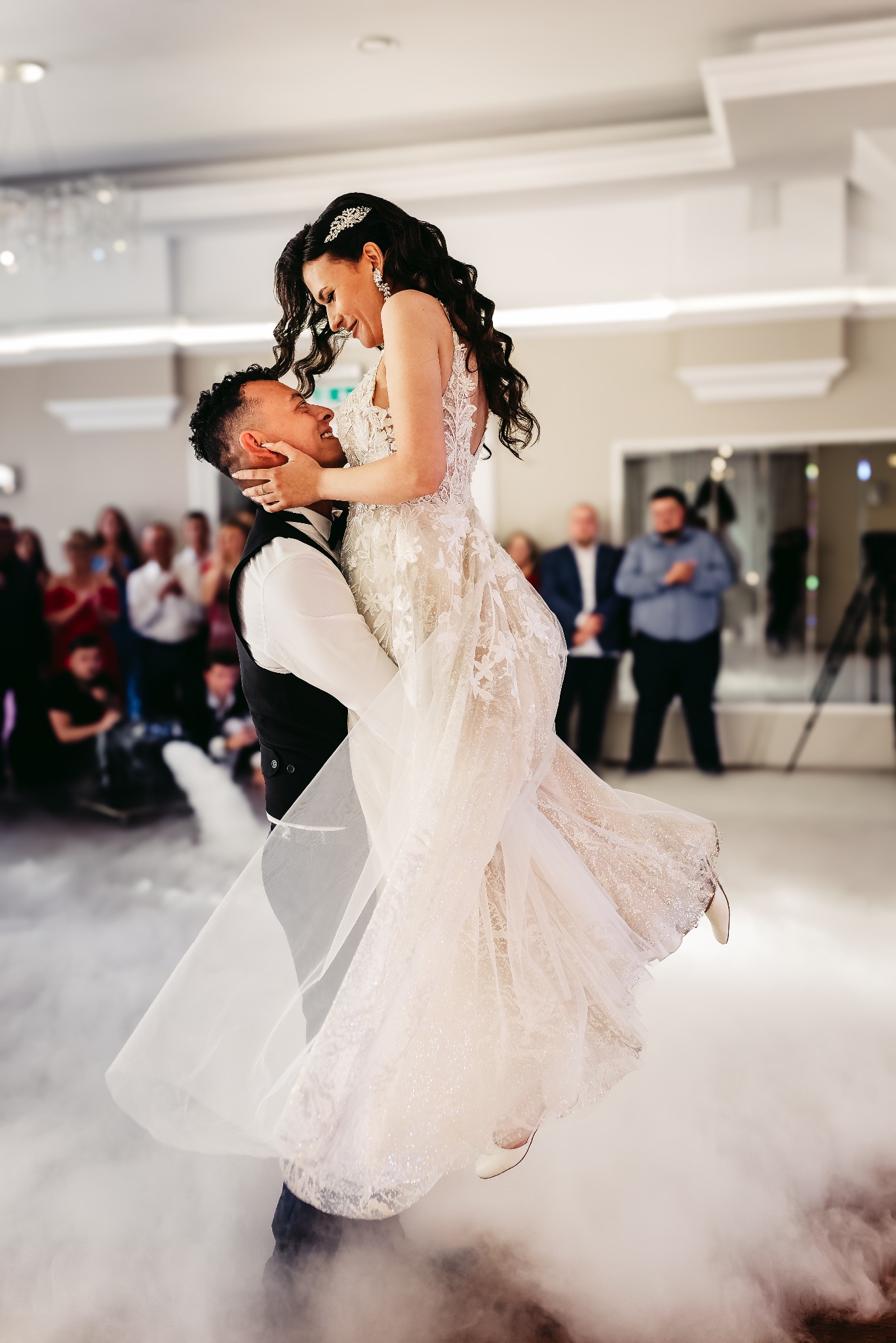 Uśmiechnięty pan młody trzyma pannę młodą w sukni z koronkowymi aplikacjami, na parkiecie unosi się mgła, w tle goście.