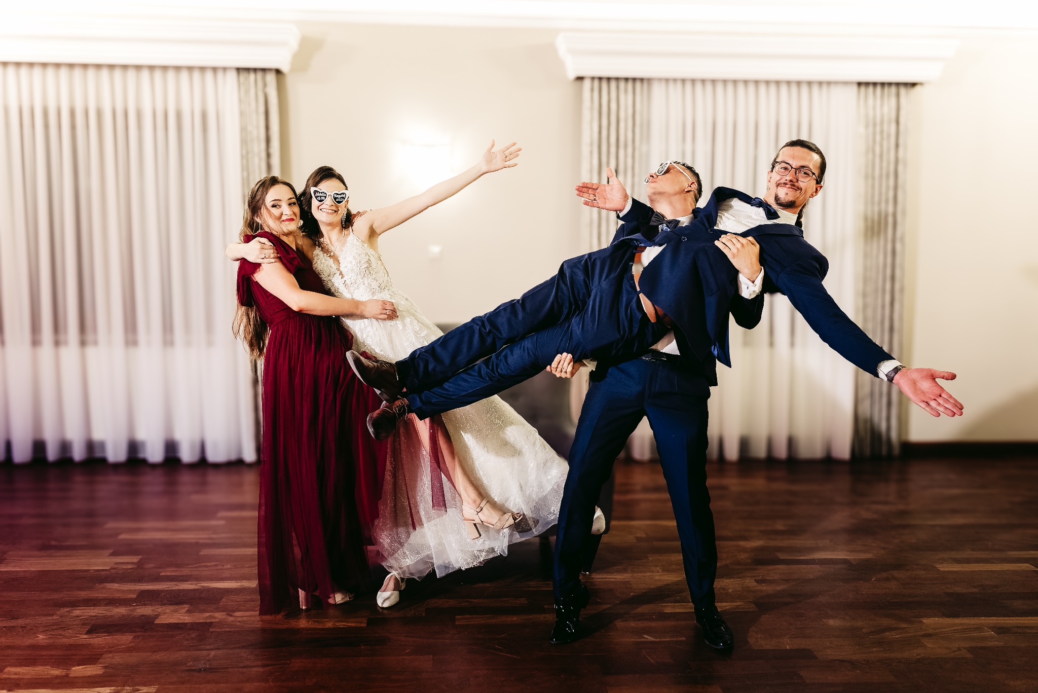 Panna młoda w białej sukni i goście w zabawnych okularach pozują, unosząc w górę mężczyznę w granatowym garniturze na parkiecie.