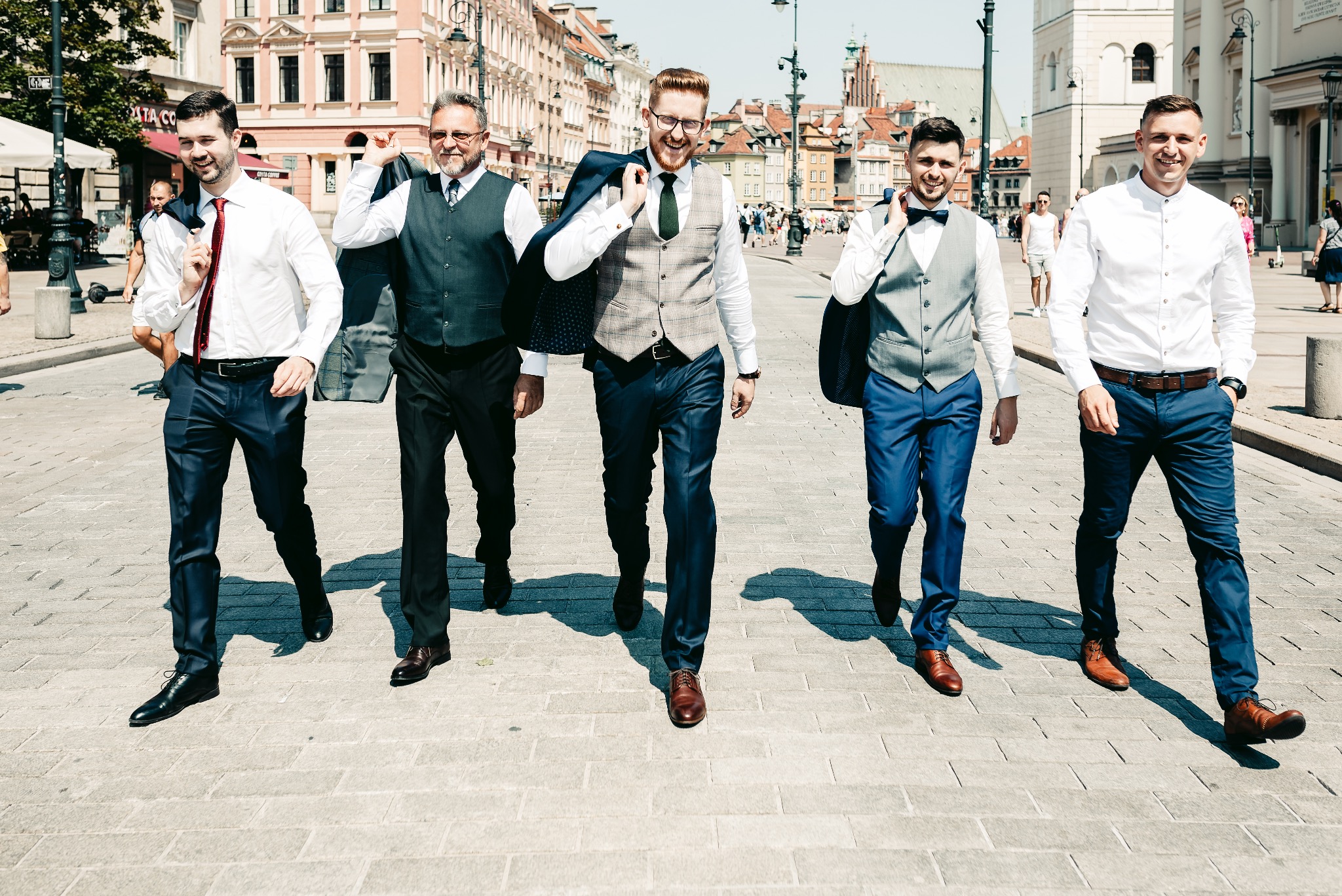 Pięciu mężczyzn w eleganckich strojach idzie po brukowanej ulicy w słoneczny dzień, niektórzy z marynarkami przewieszonymi przez ramię, w tle budynki miejskie.