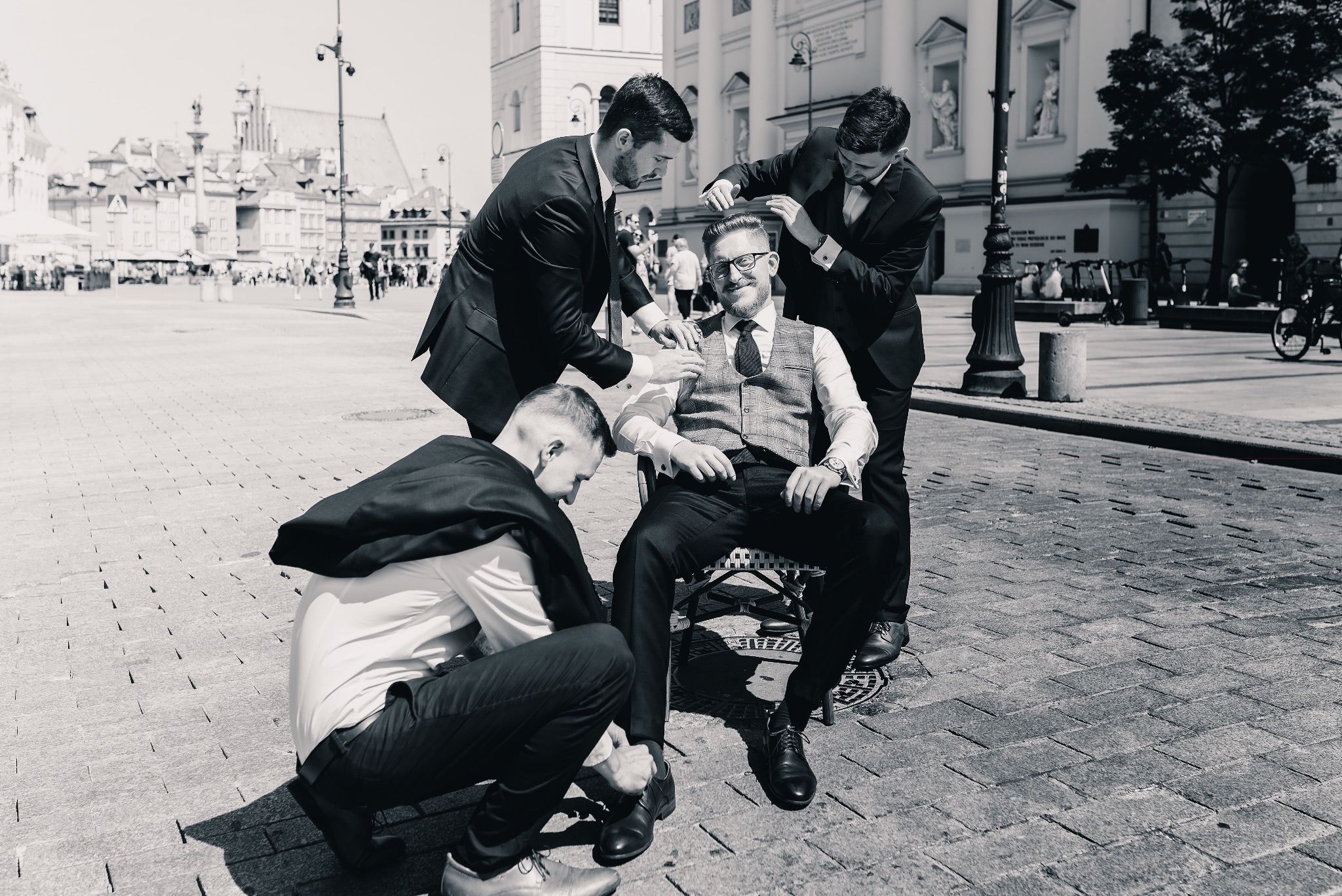 Czarno-białe zdjęcie pana młodego siedzącego na krześle, któremu poprawiają muchę i buty na tle brukowanego placu w Warszawie