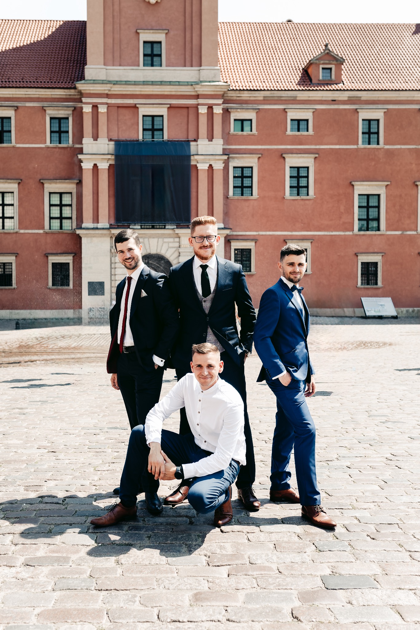 Czterech mężczyzn w garniturach pozuje na brukowanym dziedzińcu na tle budynku z czerwonej cegły z licznymi oknami.