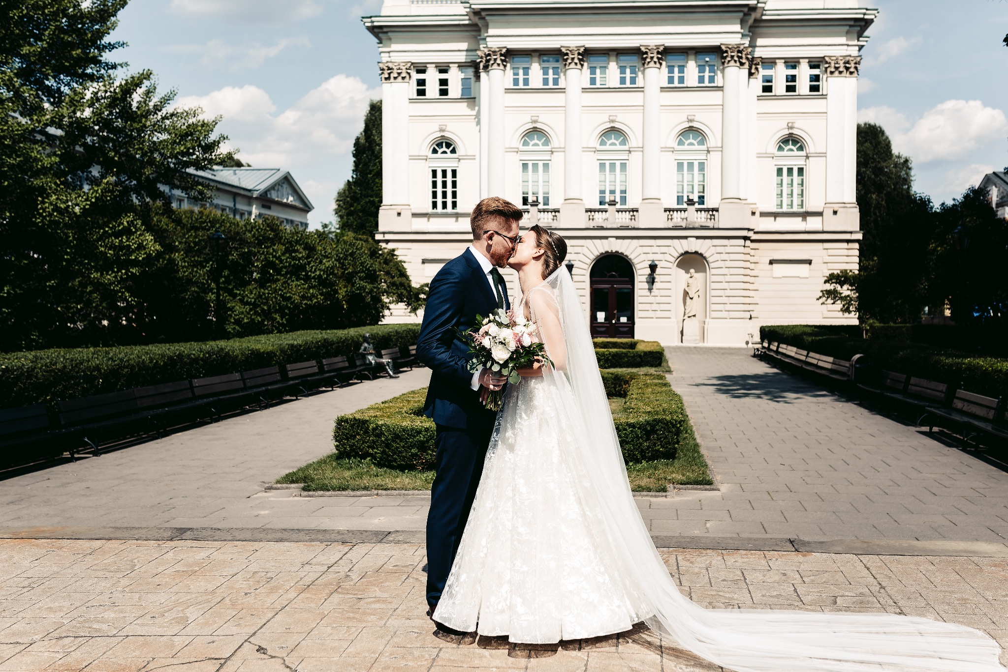 Para młoda w pocałunku na tle klasycystycznego budynku z kolumnami i zadbanym ogrodem.