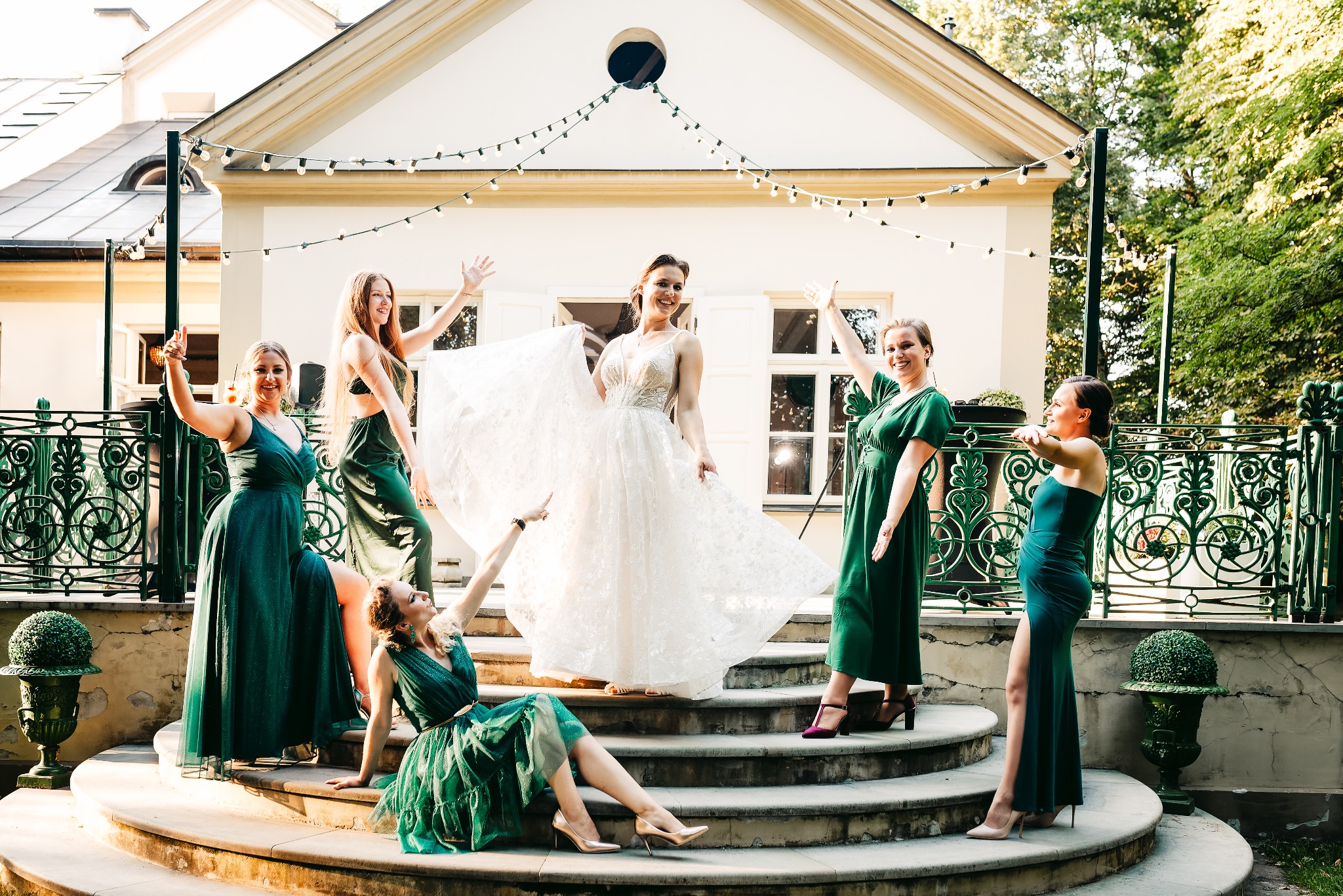 Panna młoda w koronkowej sukni i druhny w szmaragdowych sukniach pozują na schodach przed budynkiem z girlandami świetlnymi.