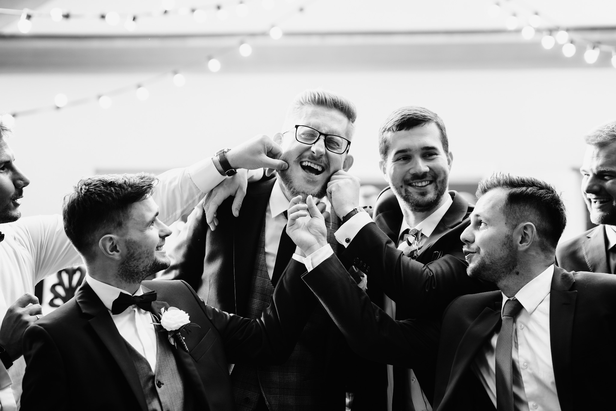 Czarno-białe zdjęcie grupy mężczyzn w garniturach, wesoło pozujących, jeden z nich ma ściskane policzki, w tle dekoracja ze światełek.