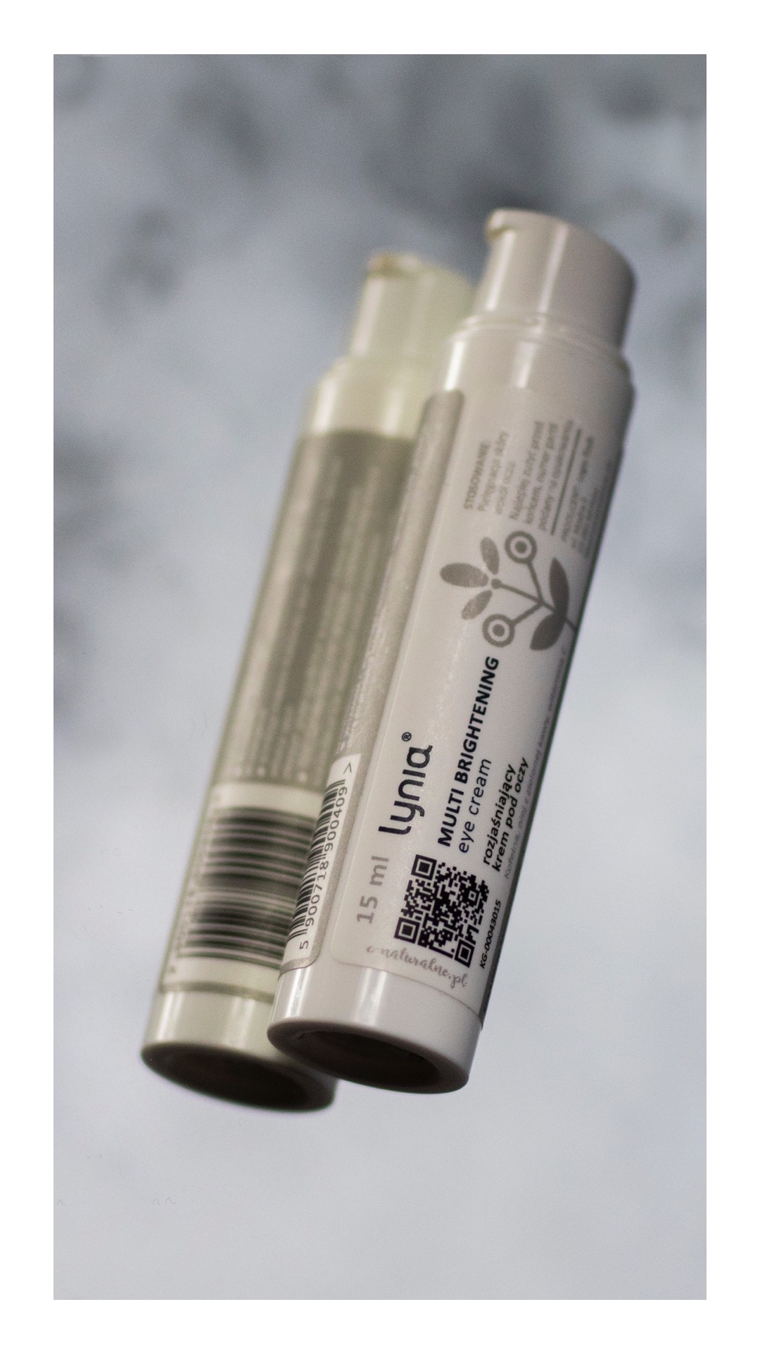 Dwie tubki kremu pod oczy Lynia Multi Brightening, jedna z kodem QR i informacjami o składzie, druga z kodem kreskowym, ustawione pod kątem na rozmytym tle.