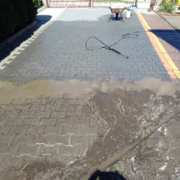 Częściowo wyczyszczona kostka brukowa na podjeździe, widoczne różnice w kolorze i zabrudzeniu, lanca myjąca leży na czystej powierzchni, taczka i wiadro w tle.