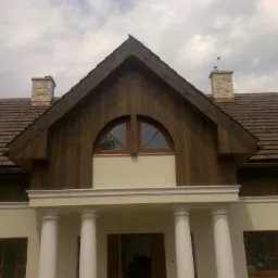 Front domu z drewnianym wykończeniem dachu, kolumnami przy wejściu i oknem w kształcie półkola na poddaszu.