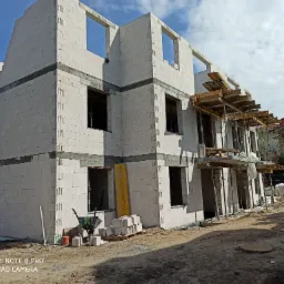 Wznoszenie dwukondygnacyjnego budynku z pustaków, widoczne otwory okienne i drzwiowe, częściowo zamontowane szalunki stropowe, taczka i materiały budowlane na placu budowy.