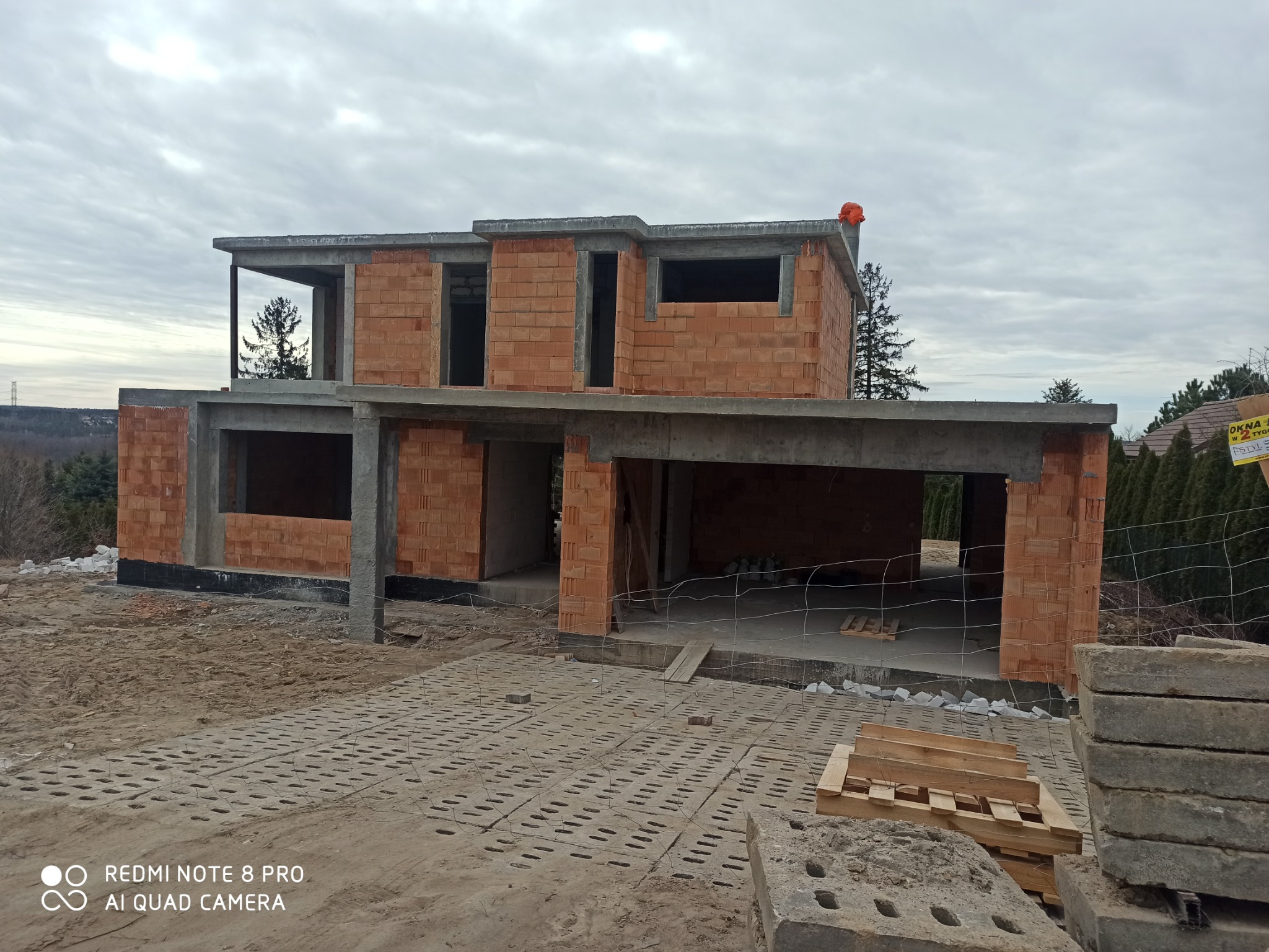 Dwukondygnacyjny dom w stanie surowym otwartym, z czerwonej cegły, widoczne betonowe stropy i nadproża, na pierwszym planie ażurowe płyty betonowe, w tle zieleń i pochmurne niebo.