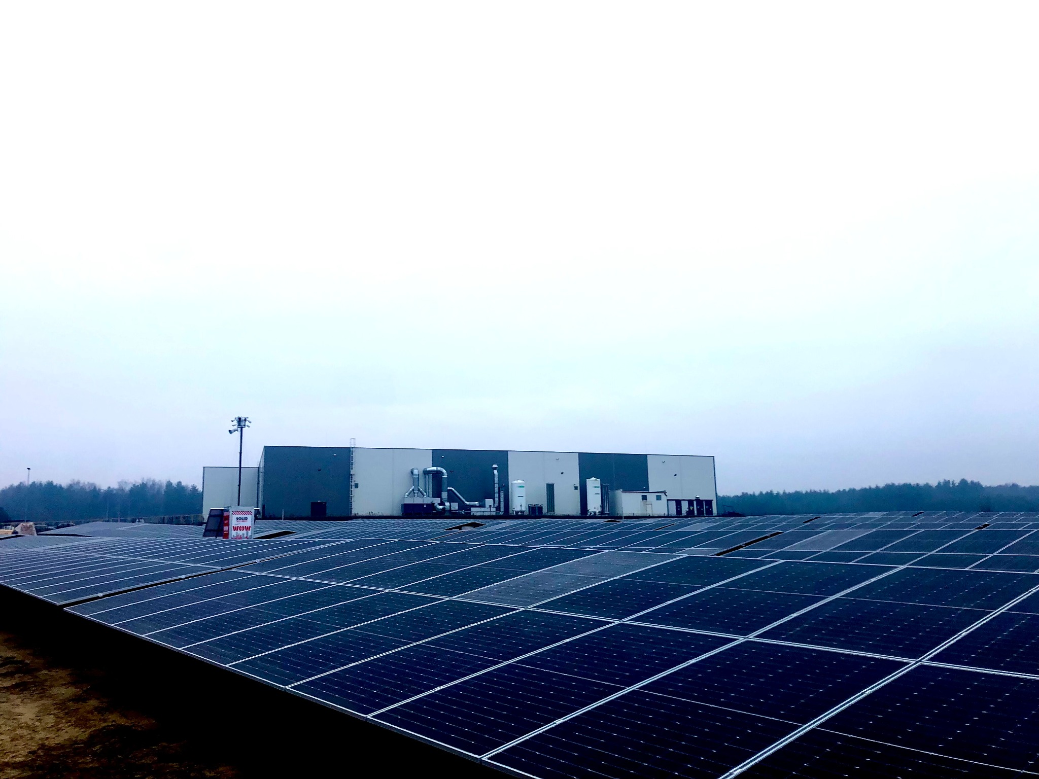 Rozległa farma paneli słonecznych z budynkiem przemysłowym w tle, widoczna tablica reklamowa na jednym z paneli, pochmurny dzień.