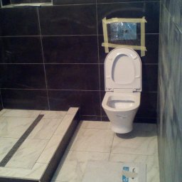 PSV-SERVICE SP.z.o.o - Nowoczesna łazienka w trakcie wykańczania: czarne płytki na ścianach, marmurowe płytki na podłodze z odpływem liniowym i wisząca toaleta z otwartą klapą. Na ścianie zabezpieczone taśmą malarską...