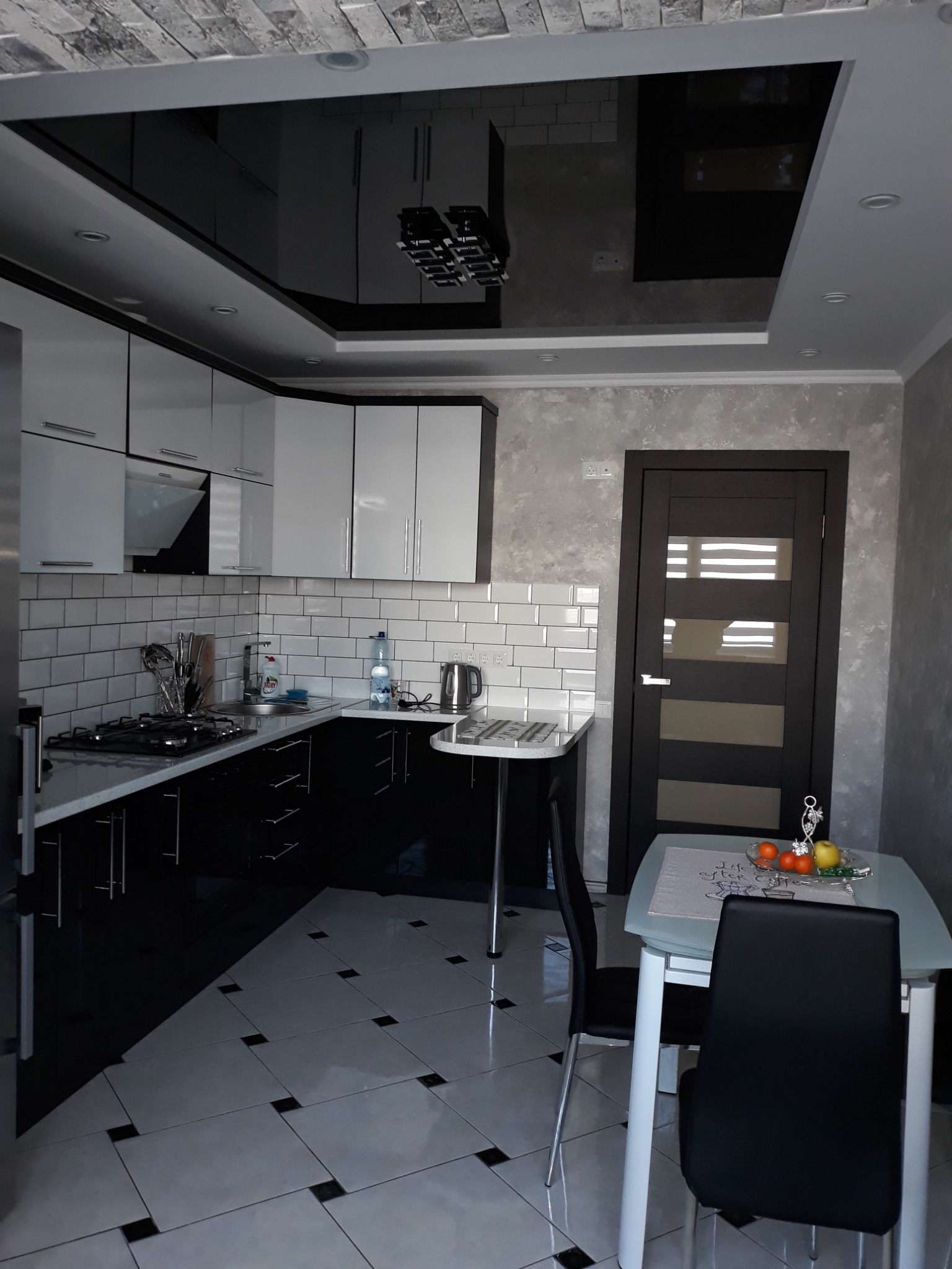 Nowoczesna kuchnia w czarno-białej kolorystyce z błyszczącym, czarnym sufitem podwieszanym i białymi płytkami na ścianie, uzupełniona stolikiem z owocami.