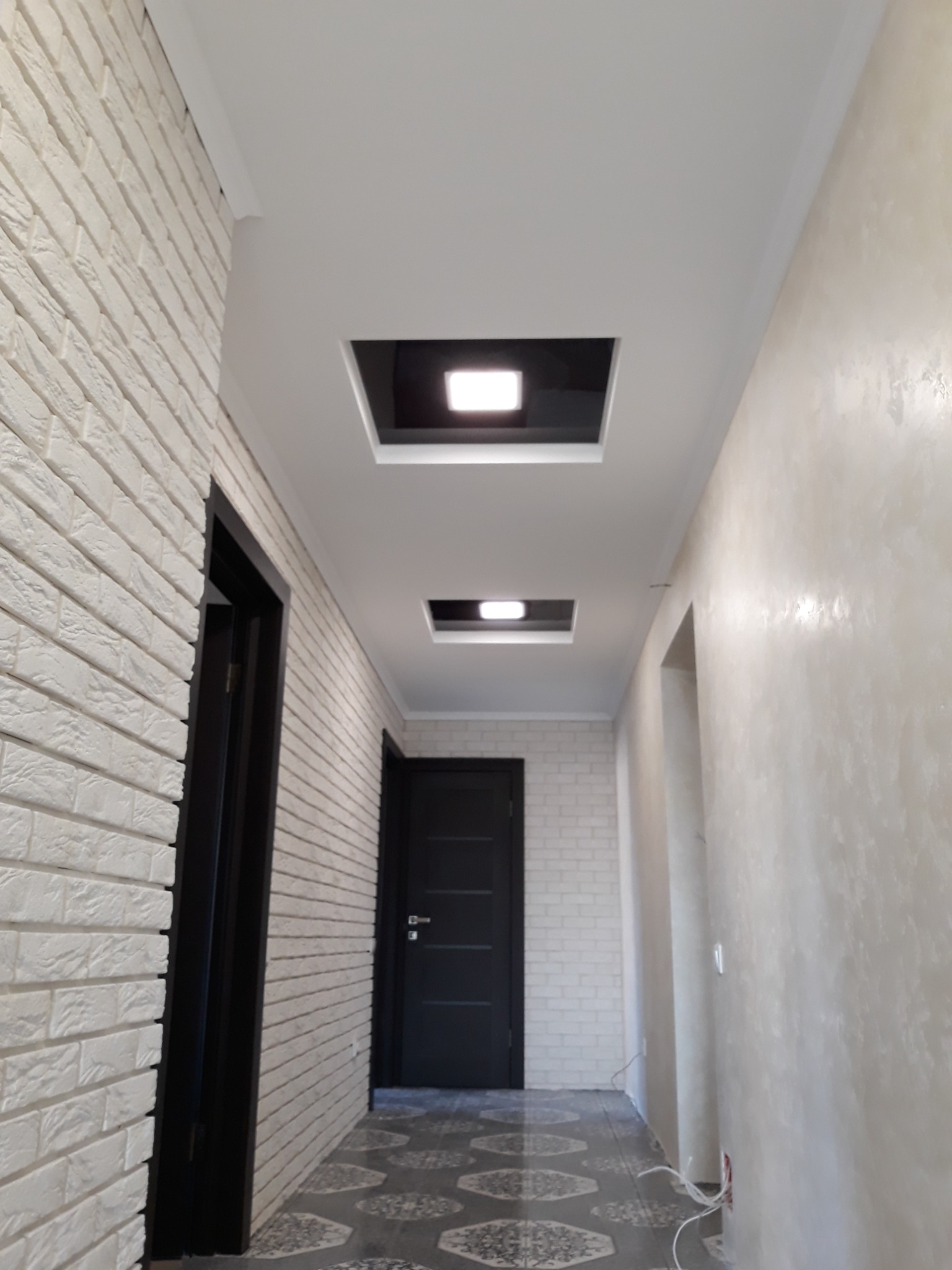 Korytarz po remoncie: ściana z białej cegły, ściana z efektem dekoracyjnym, sufit z wpuszczonymi kwadratowymi lampami, ciemne drzwi i wzorzysta podłoga.