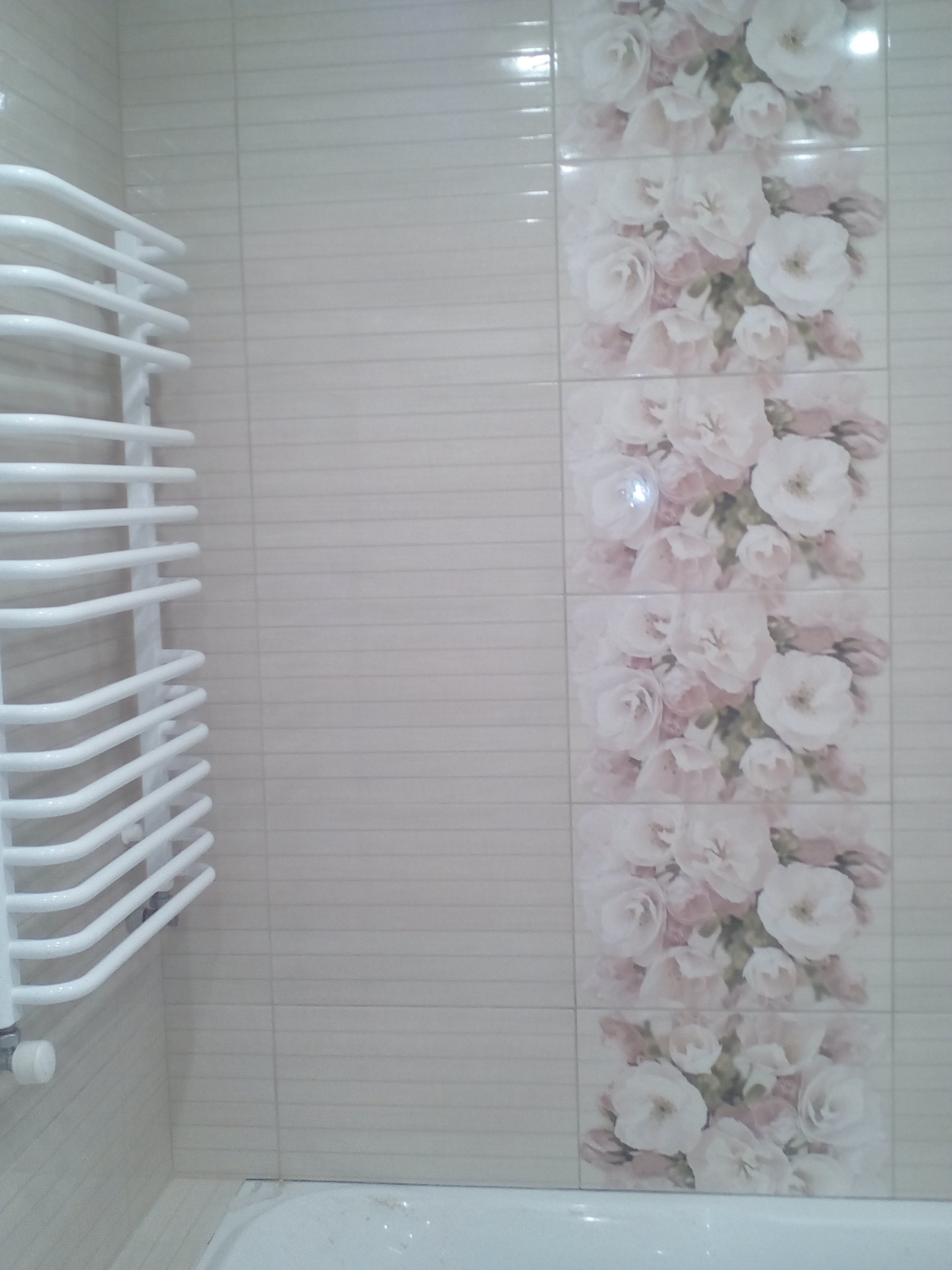 Wykończona ściana łazienki z beżowymi płytkami i pionowym pasem płytek dekoracyjnych z motywem białych i różowych kwiatów, obok biały grzejnik drabinkowy.