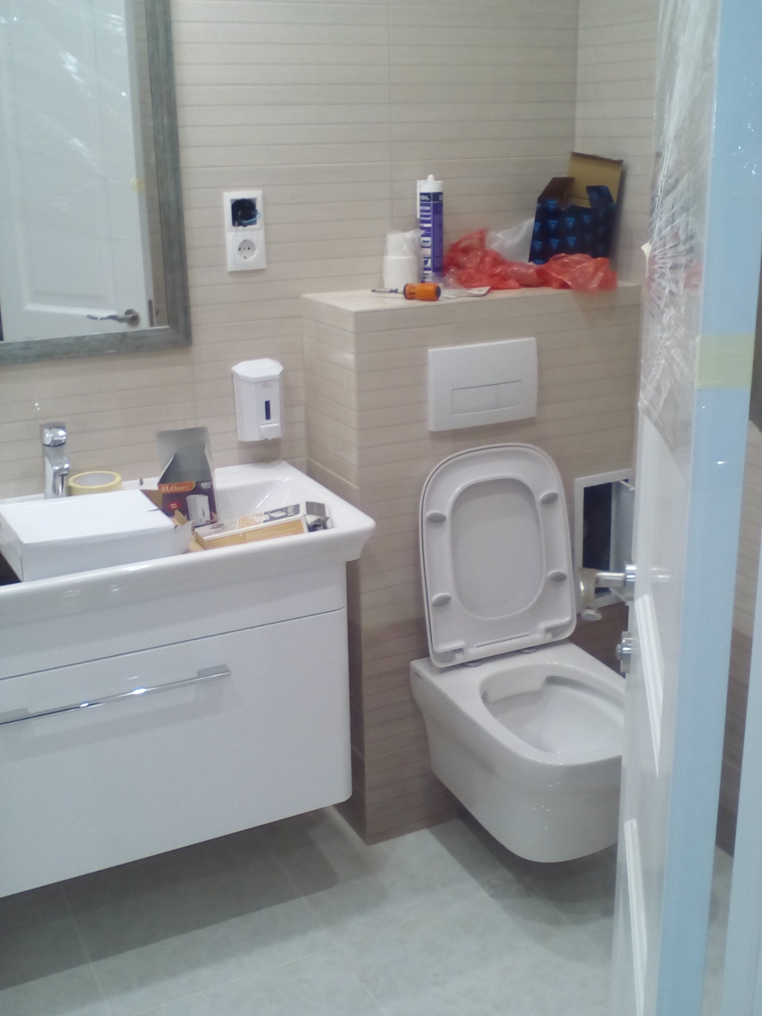 Nowa, biała toaleta podwieszana z otwartą klapą, umywalka z szafką i lustro w łazience z beżowymi płytkami, widoczne materiały budowlane i narzędzia sugerują prace wykończeniowe.