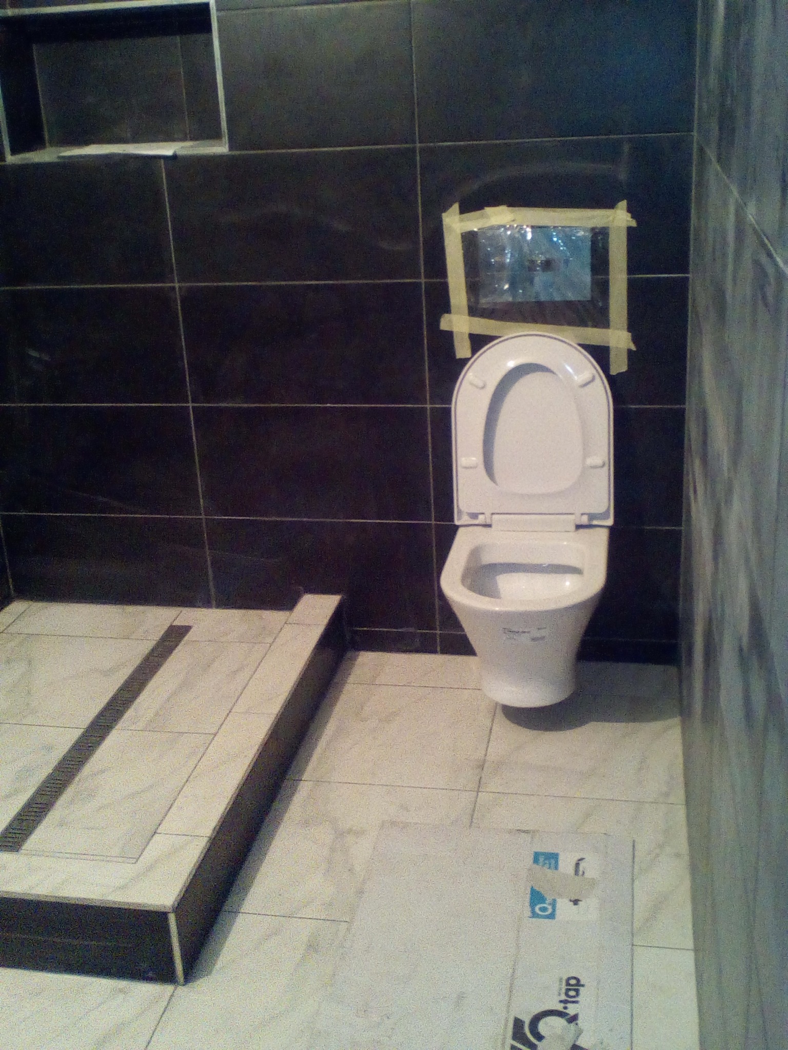 Nowoczesna łazienka w trakcie wykańczania: czarne płytki na ścianach, marmurowe płytki na podłodze z odpływem liniowym i wisząca toaleta z otwartą klapą. Na ścianie zabezpieczone taśmą malarską...