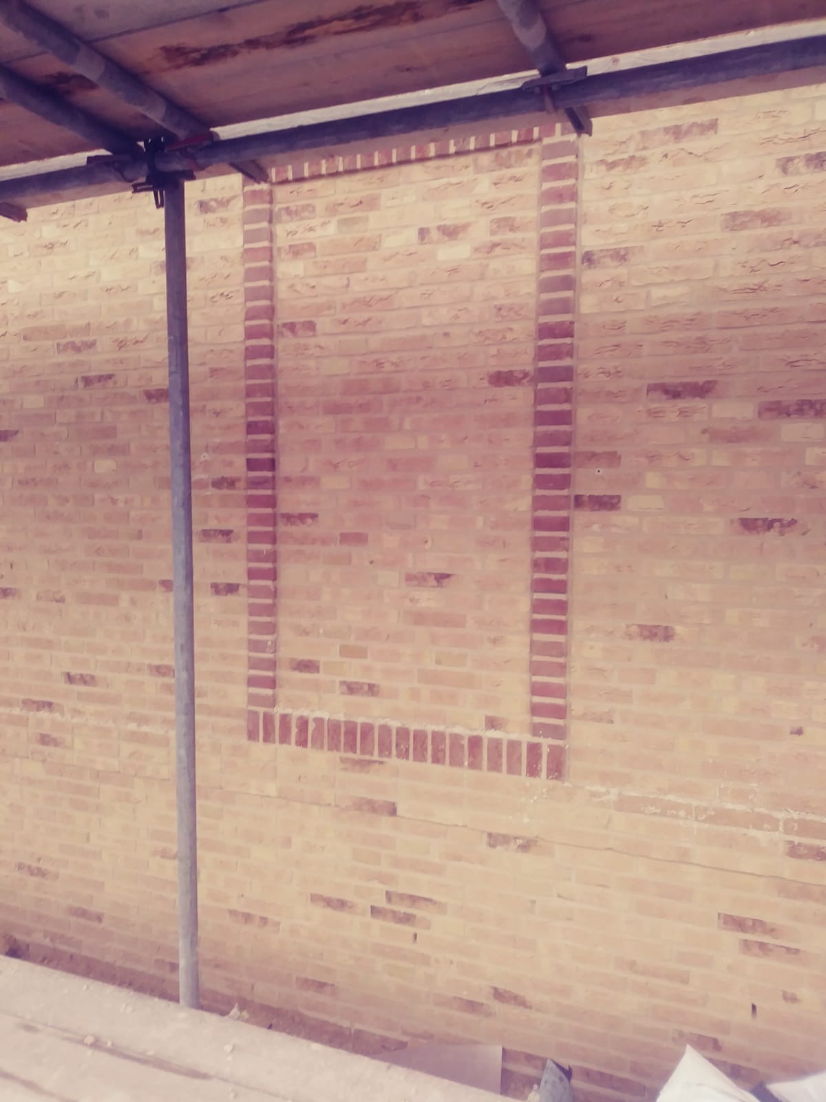 Wznoszona ściana z cegły z zaznaczonym otworem okiennym, widoczne rusztowanie i jasna kolorystyka cegieł.