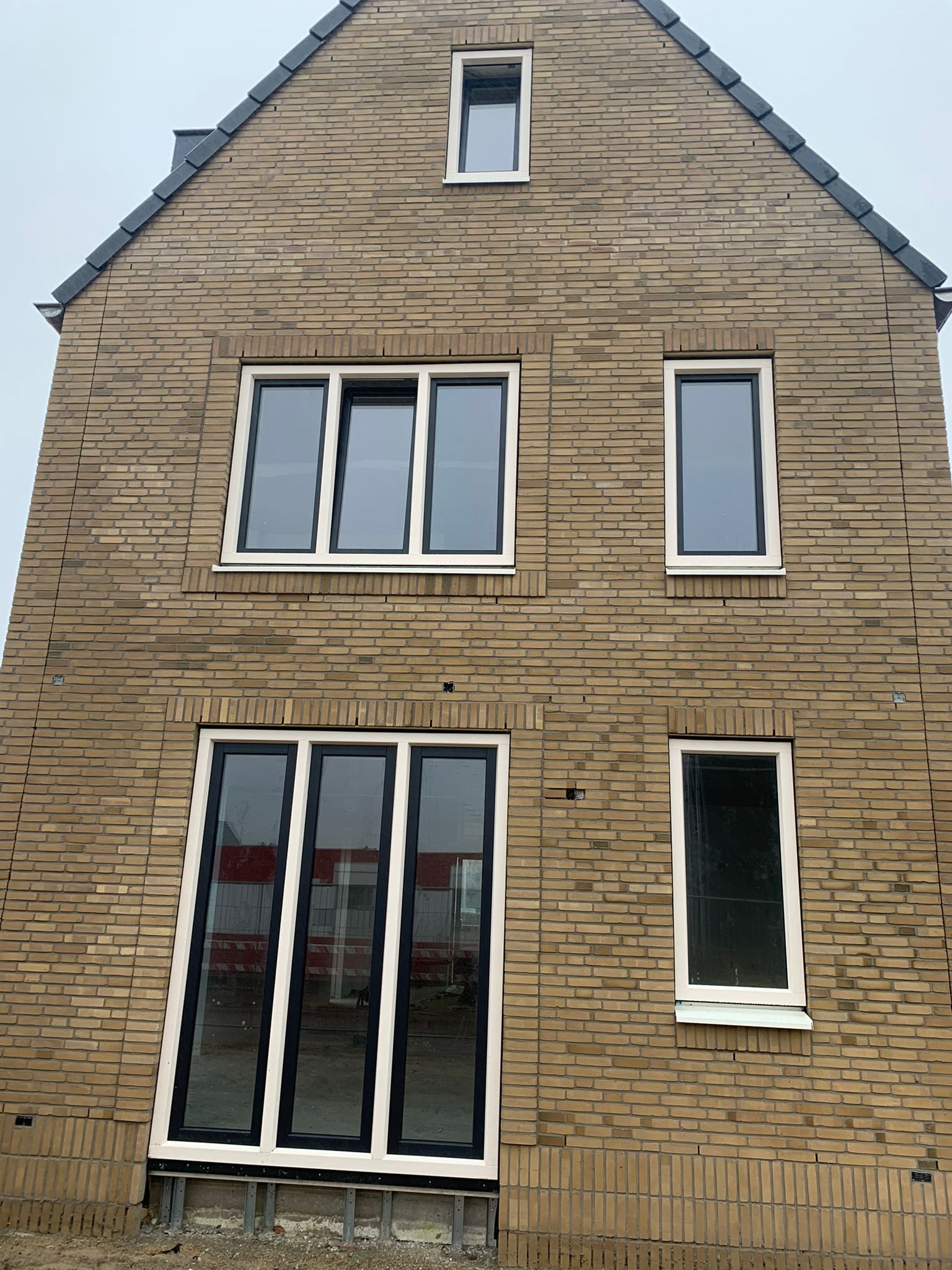 Dwukondygnacyjny dom w trakcie budowy, z widoczną elewacją z żółtej cegły, czarnymi ramami okiennymi z białymi obramowaniami i konstrukcją fundamentową w dolnej części.