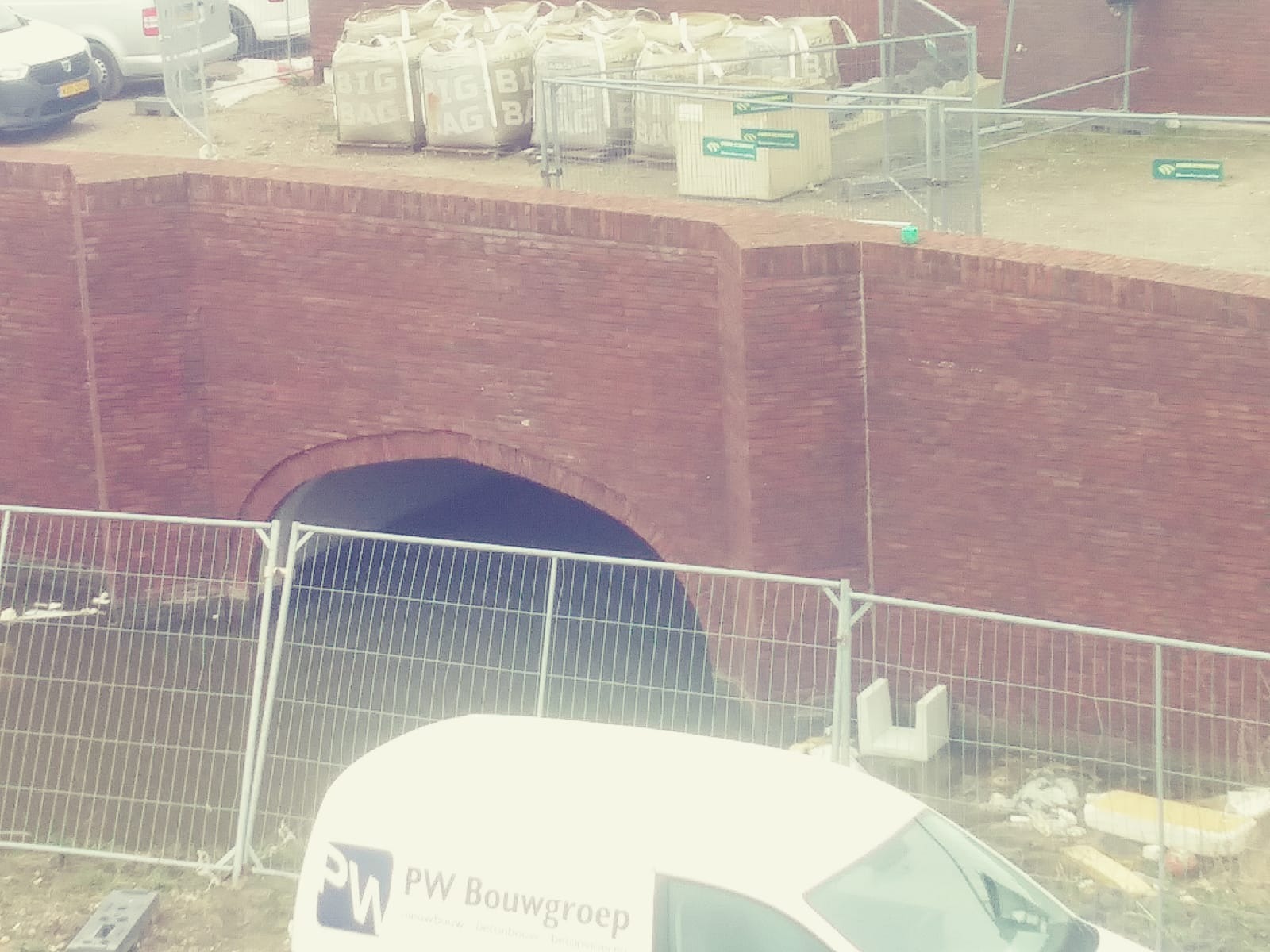 Ceglany mur oporowy z tunelem w trakcie budowy, widoczne worki z materiałami budowlanymi, ogrodzenie tymczasowe i samochód dostawczy.