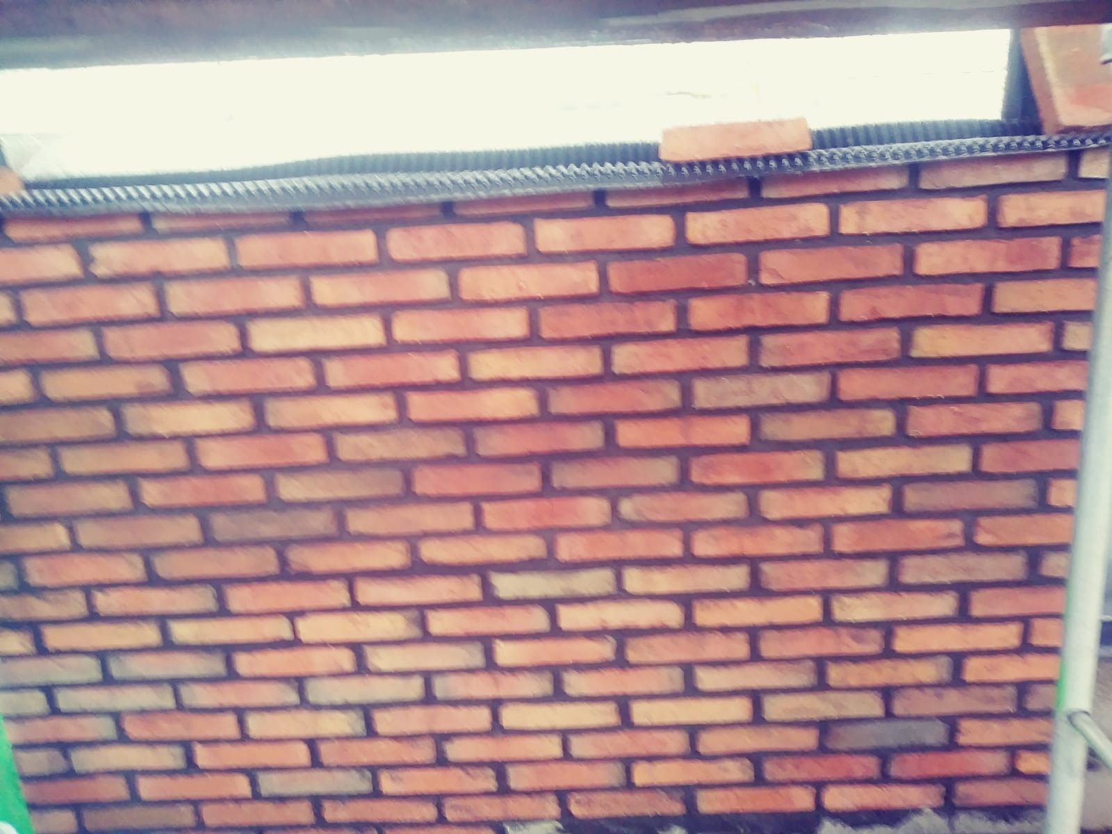 Świeżo wymurowana ściana z czerwonej cegły, widoczne czarne spoiny, u góry element konstrukcyjny z czarną folią i pojedynczą cegłą, widok z dołu.