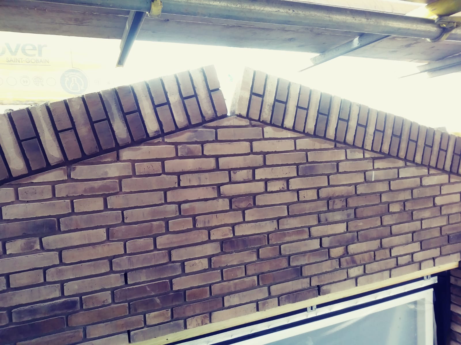 Wznoszenie ściany z cegły klinkierowej w kształcie szczytu, widoczne rusztowanie i fragment okna, etap budowy.