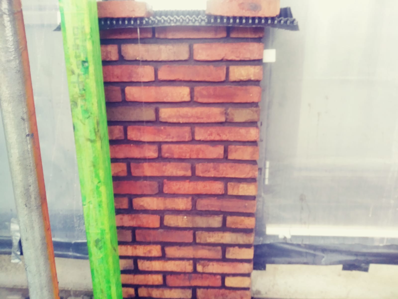 Fragment nowo wzniesionej ściany z czerwonej cegły, widoczne elementy rusztowania i folia ochronna.
