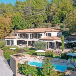 Dwupiętrowa willa z basenem na wzgórzu, otoczona bujną zielenią, z widocznymi leżakami i parasolami, oraz fragmentem drogi dojazdowej.