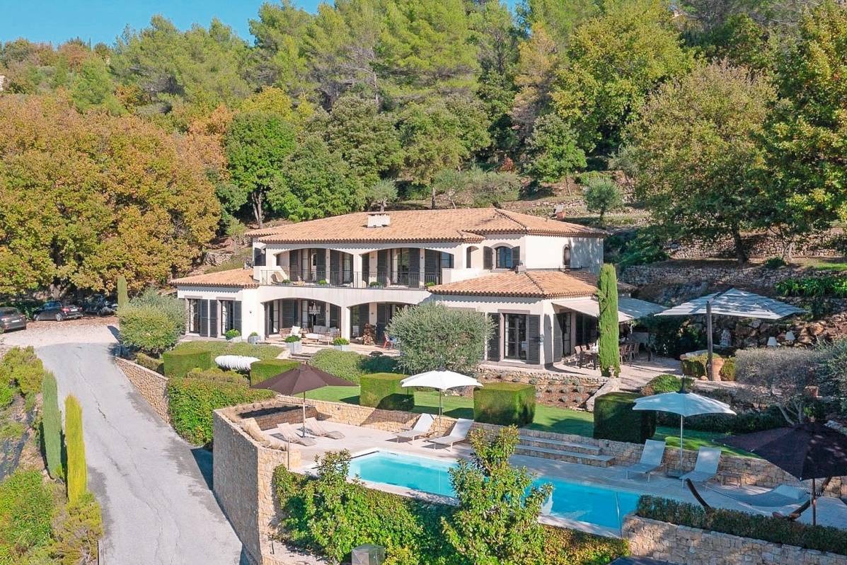 Dwupiętrowa willa z basenem na wzgórzu, otoczona bujną zielenią, z widocznymi leżakami i parasolami, oraz fragmentem drogi dojazdowej.