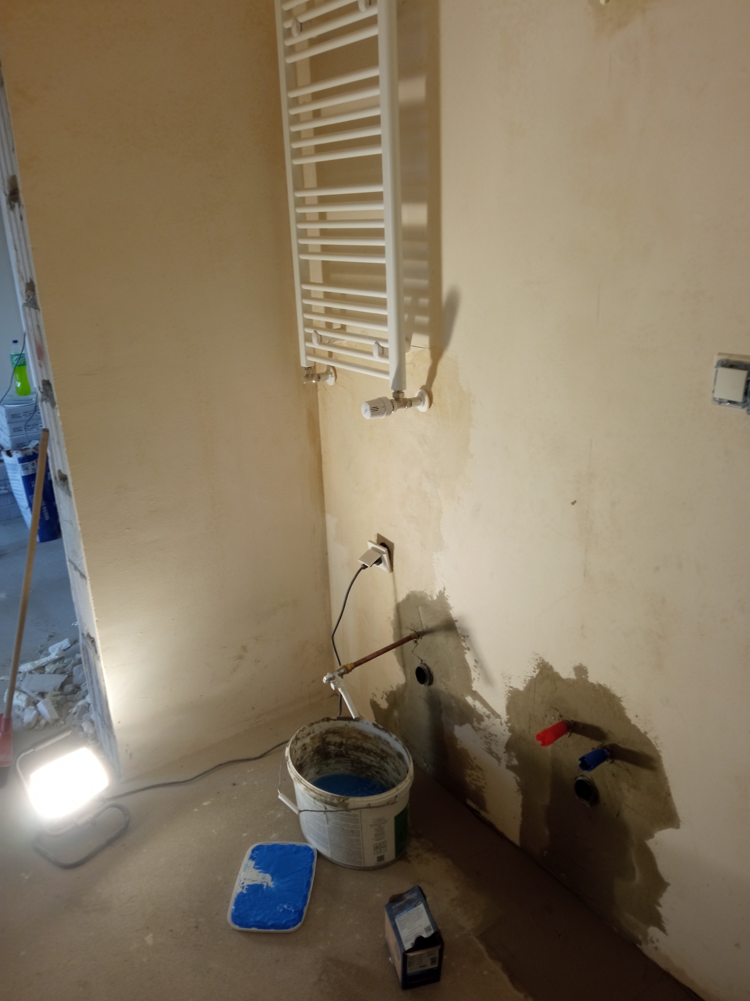 Przygotowanie ściany w łazience przed położeniem płytek, widoczna instalacja wodna z zaślepkami, wiadro z niebieską substancją, lampa budowlana oświetlająca podłogę.