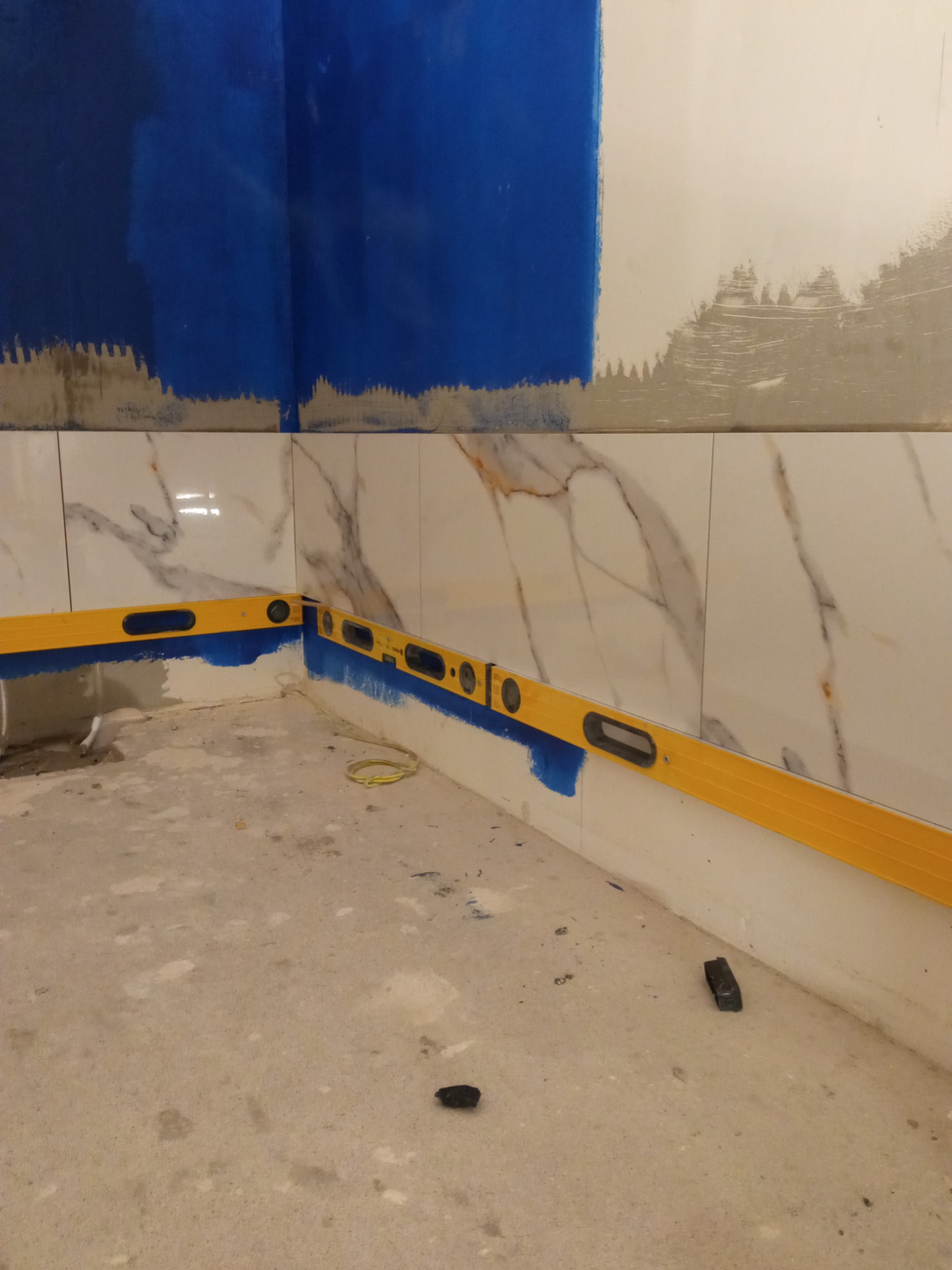 Fragment łazienki w trakcie prac: położone płytki imitujące marmur zabezpieczone niebieską taśmą malarską, żółta poziomica narożna, ślady farby na ścianie i surowa posadzka.